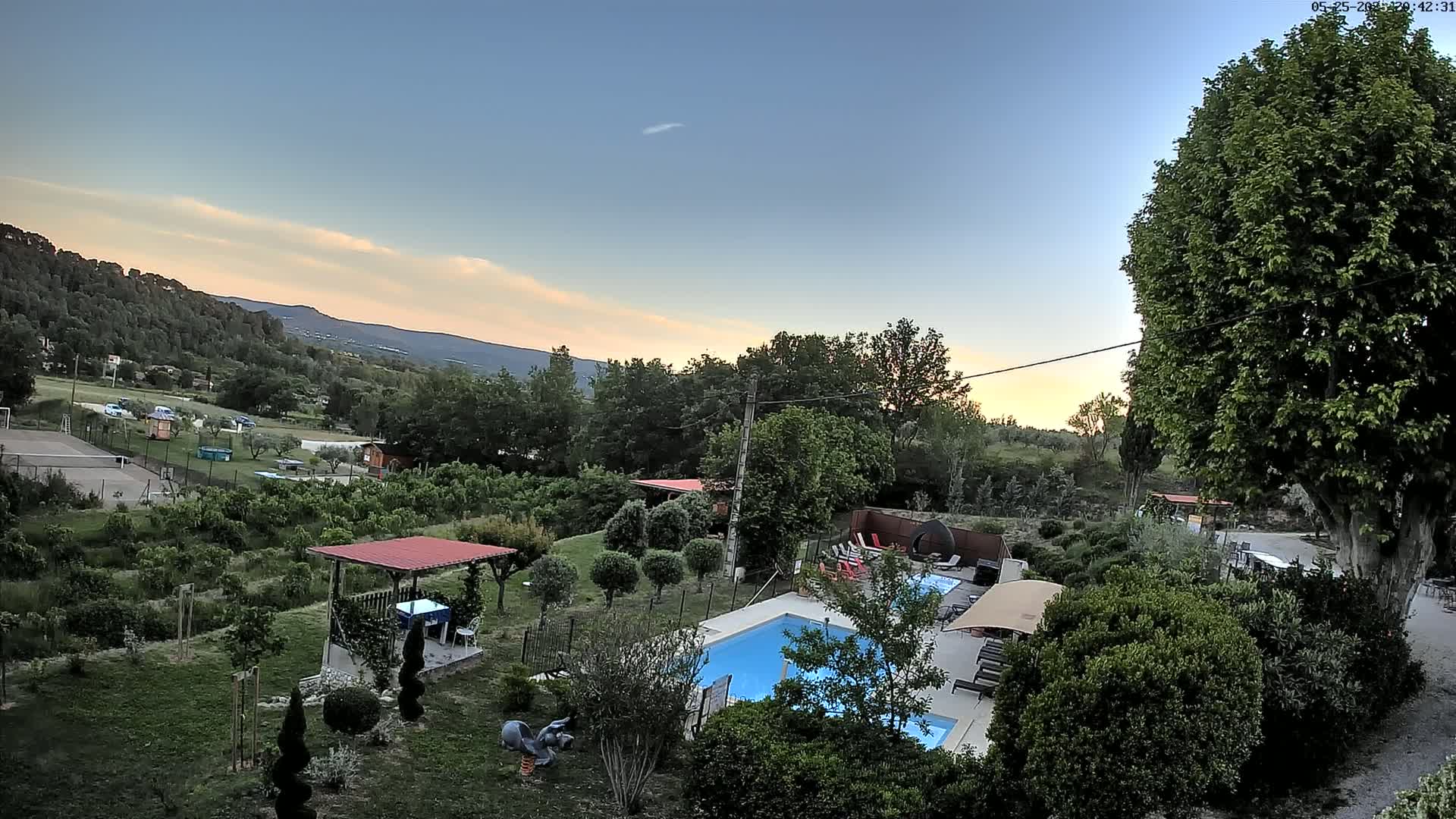 An aerial view shows a swimming pool and surrounding garden area nestled in a verdant landscape under a twilight sky.