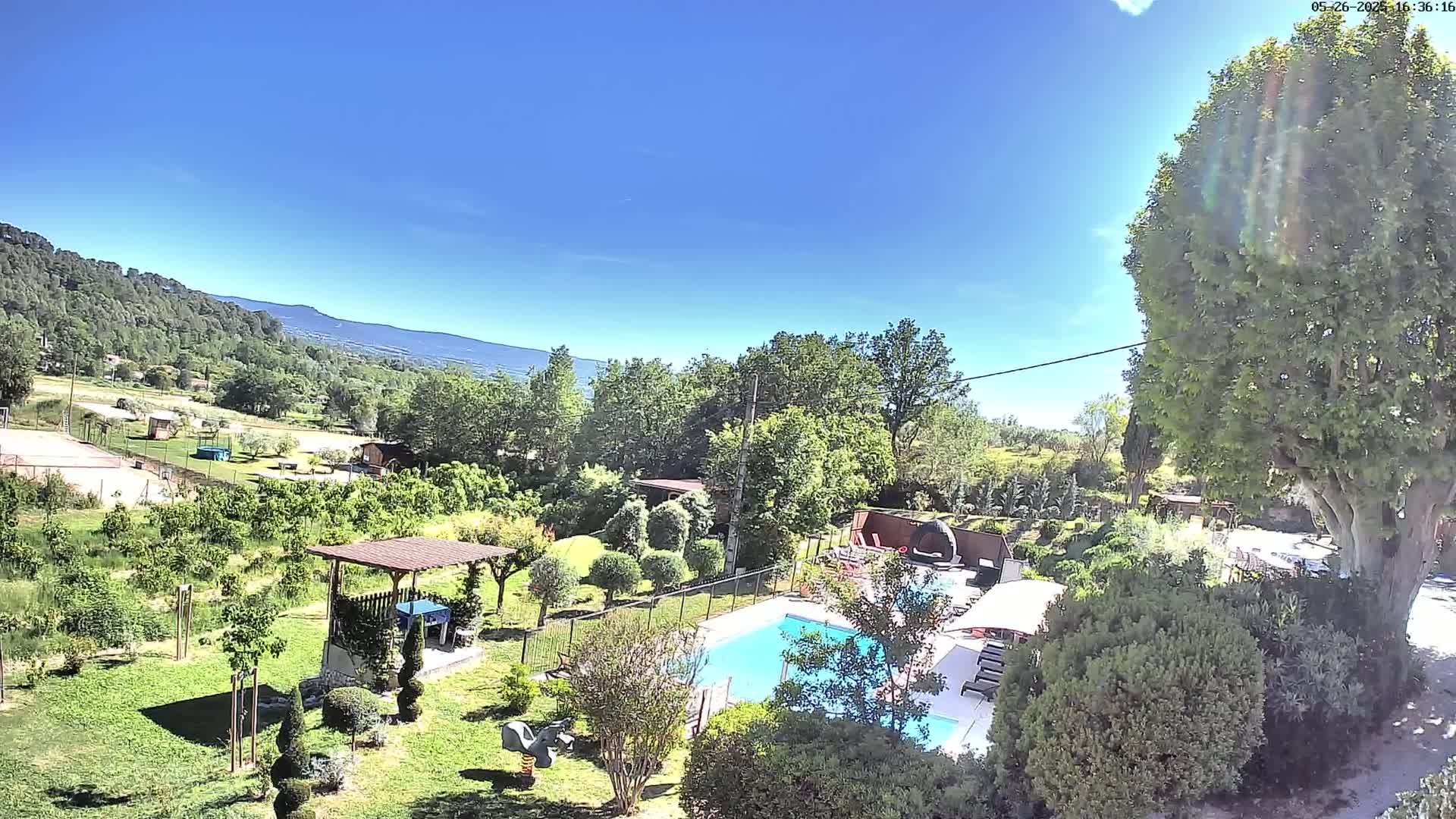 An aerial view shows a swimming pool and surrounding garden in a sunny, rural setting with a mountain range in the distance.