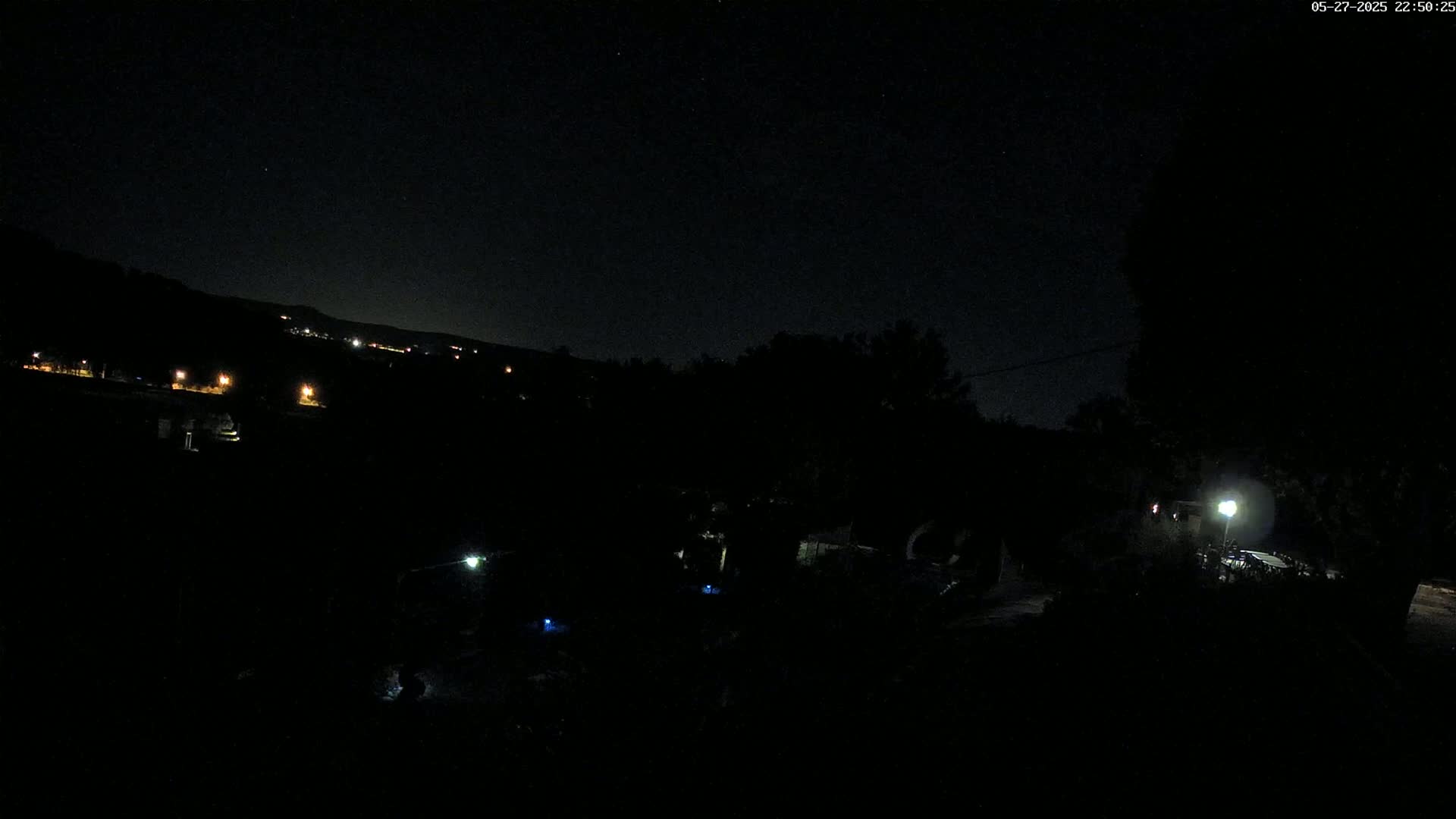 A nighttime, outdoor view shows a dark landscape with a few distant lights, a hill in the background, and a single illuminated area in the foreground.