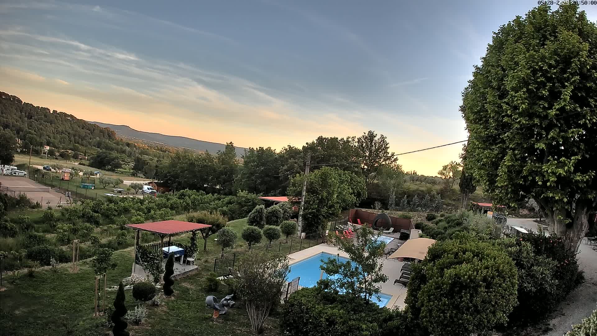 An aerial view of a residential swimming pool and surrounding garden, nestled amongst lush greenery and overlooking a mountainous landscape under a partly cloudy sunset sky.