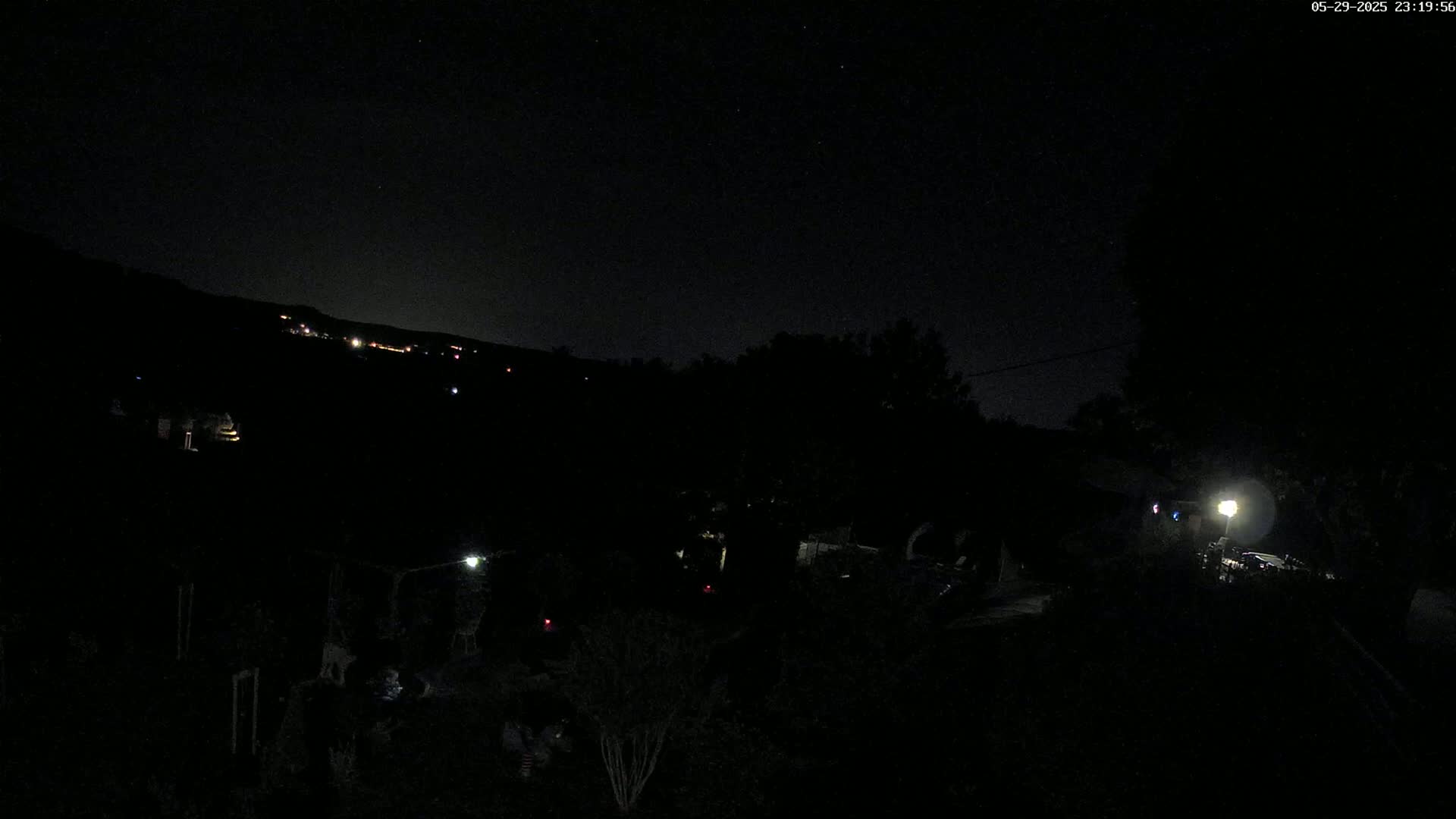 A nighttime view of a dark landscape with scattered lights, a hill in the distance, and a bright light source near the bottom right corner.