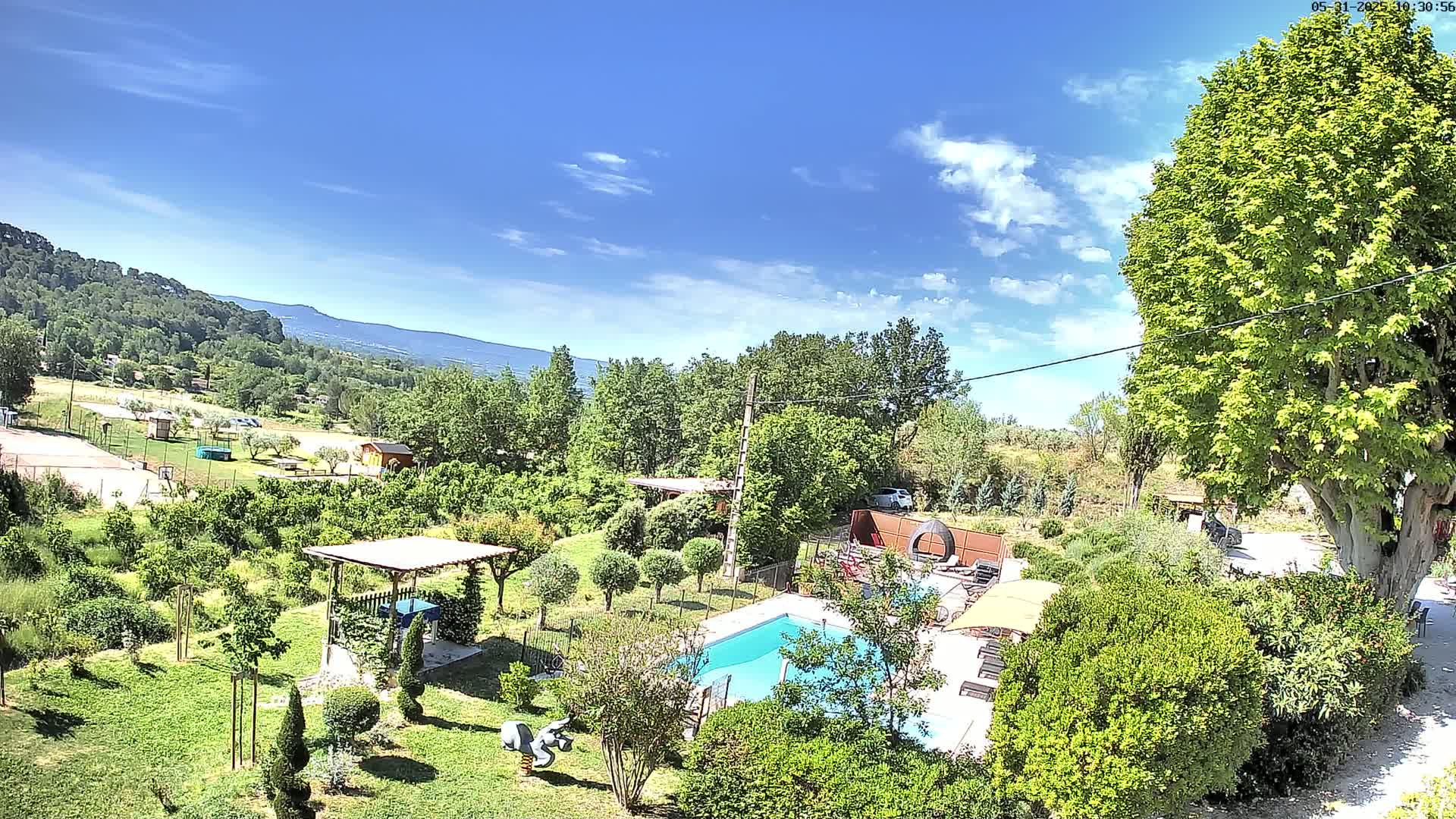 A swimming pool is nestled in a lush, green garden overlooking a hilly landscape under a mostly sunny sky with a few scattered clouds.