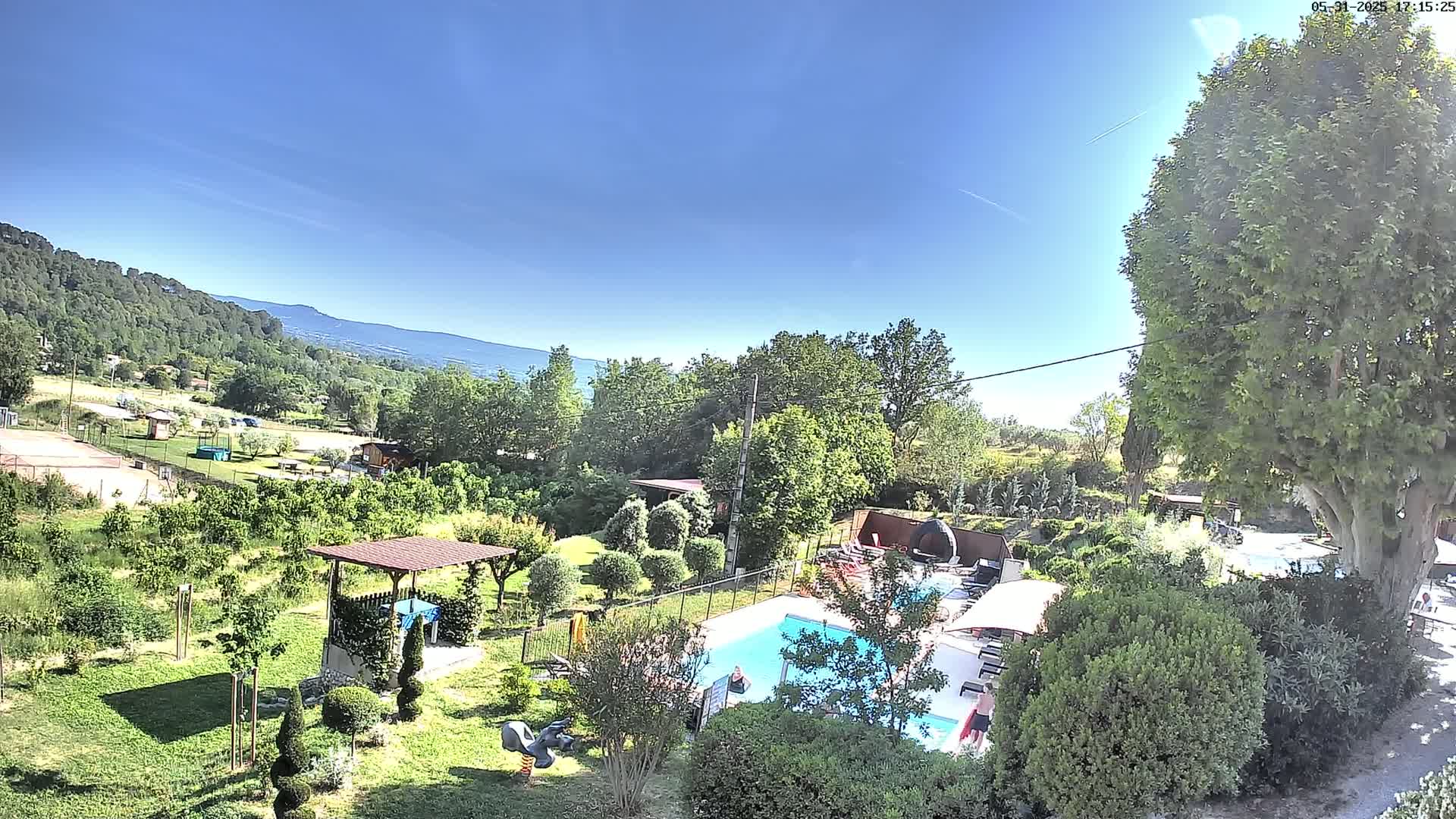 A swimming pool sits in a lush, green landscape under a clear blue sky, with a mountain range visible in the distance.