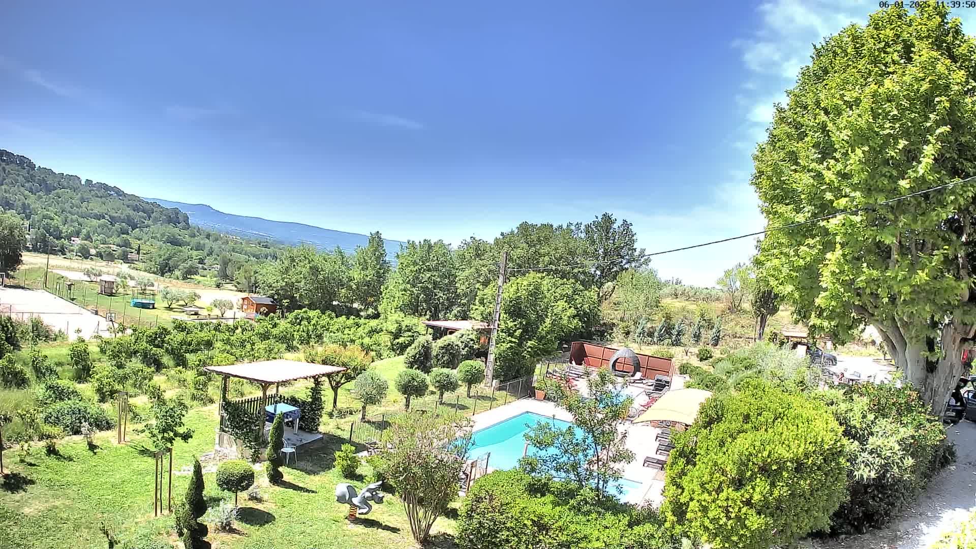 An aerial view of a swimming pool and surrounding gardens, on a sunny day with a mountain range visible in the background.
