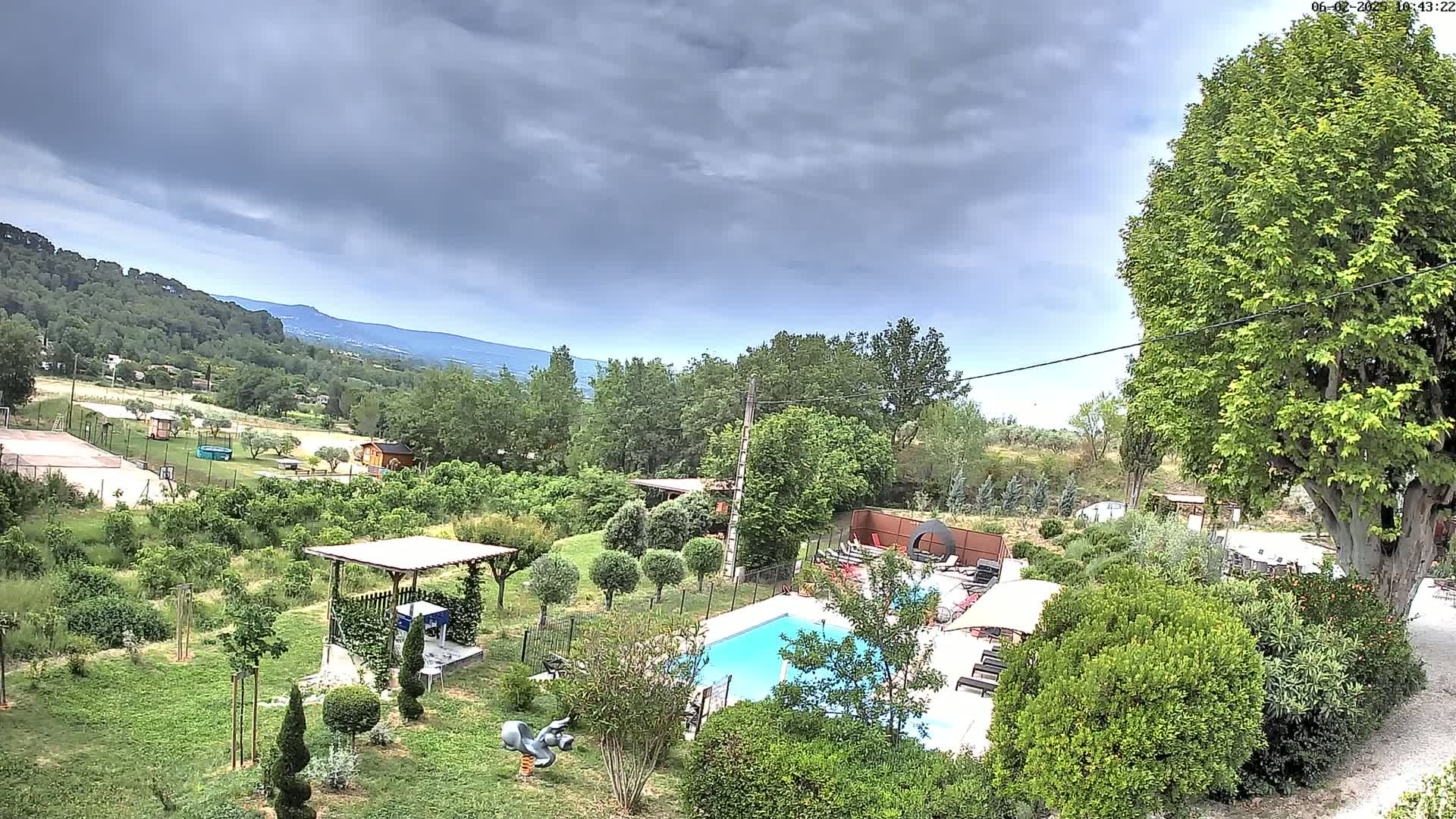 An aerial view of a swimming pool and surrounding garden, nestled amongst lush greenery and overlooking a hilly landscape under a cloudy sky.