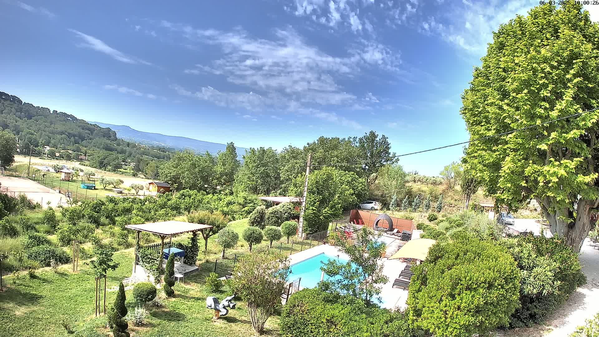 An aerial view shows a swimming pool and surrounding garden with lush greenery, a pergola, and distant mountains under a partly cloudy blue sky.
