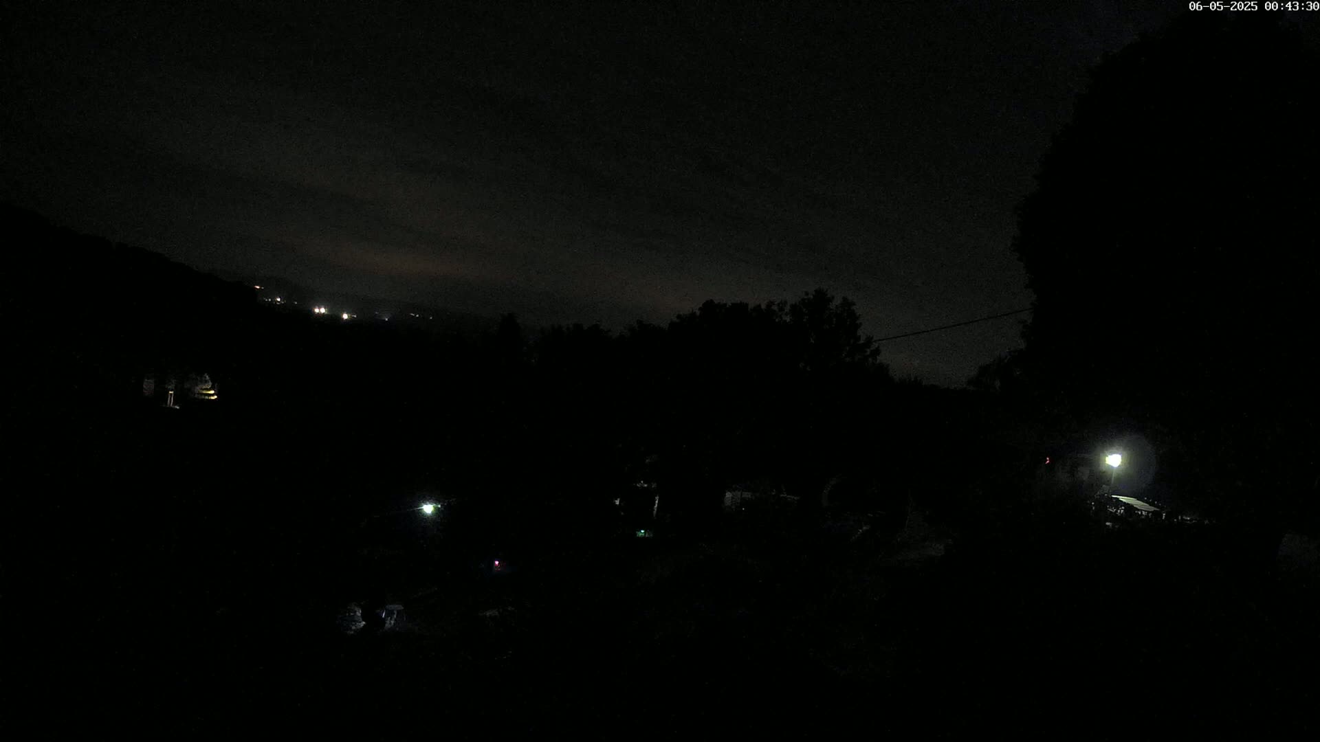 A dark nighttime scene shows a hilly landscape with sparse, dimly lit areas and trees under a mostly cloudy sky.