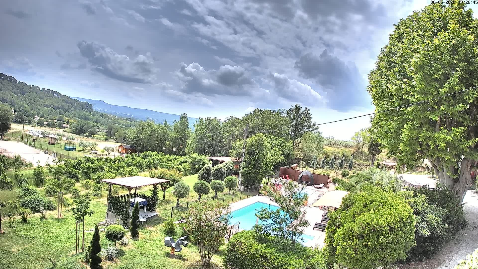 An aerial view shows a swimming pool and surrounding lush green gardens, under a partly cloudy sky, with hills and trees in the distance.