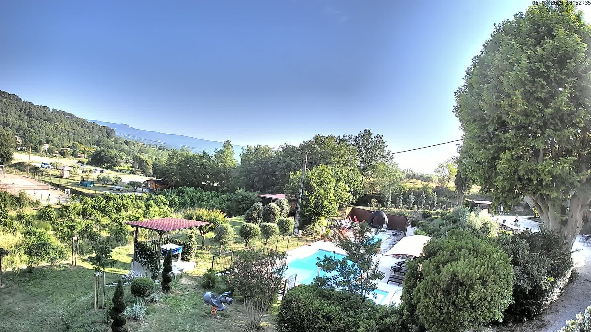 A swimming pool surrounded by a lush green garden and trees, with a view of distant mountains under a clear blue sky.
