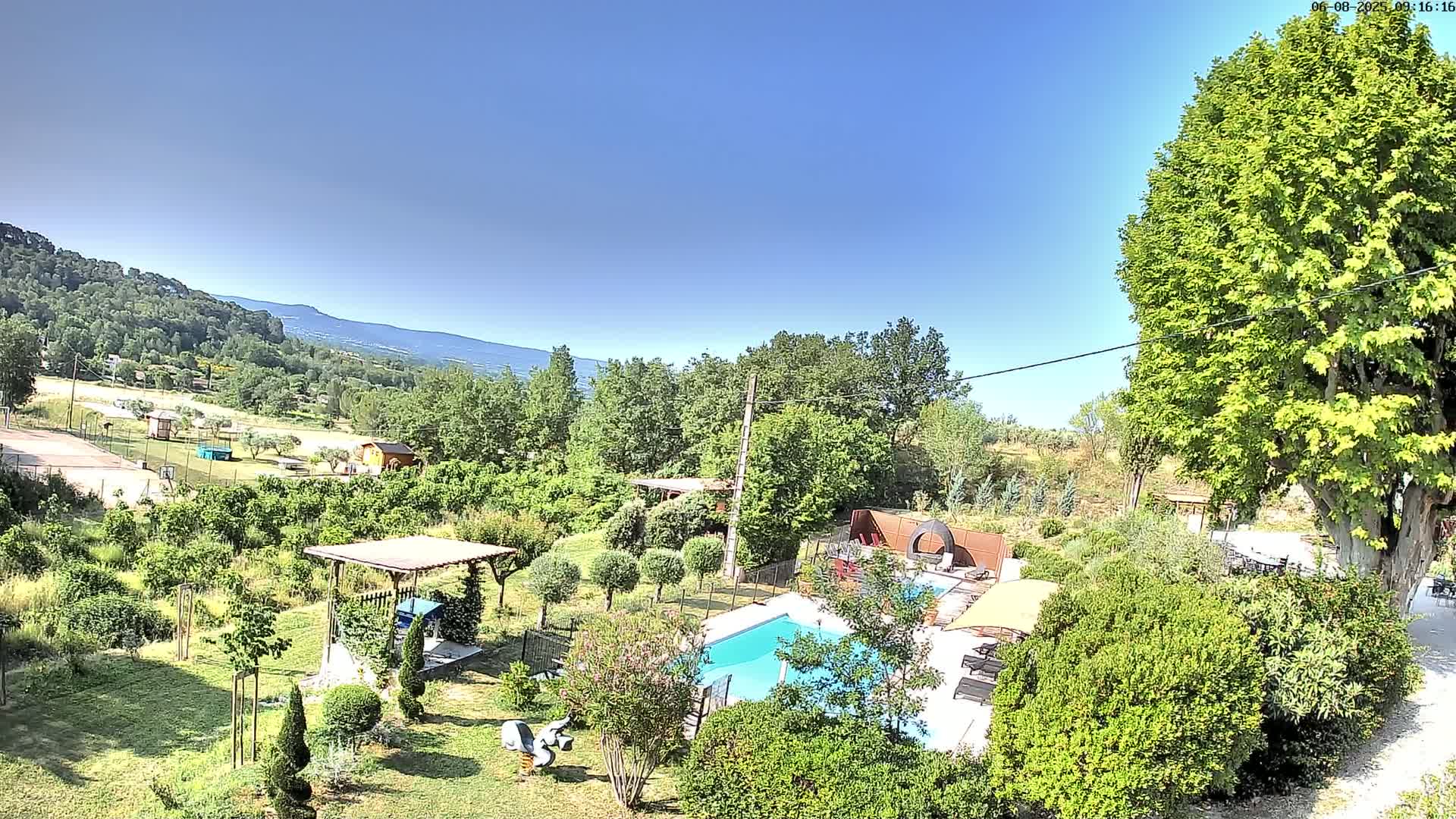 An aerial view shows a swimming pool and lush gardens in a rural setting on a clear, sunny day, with mountains visible in the distance.