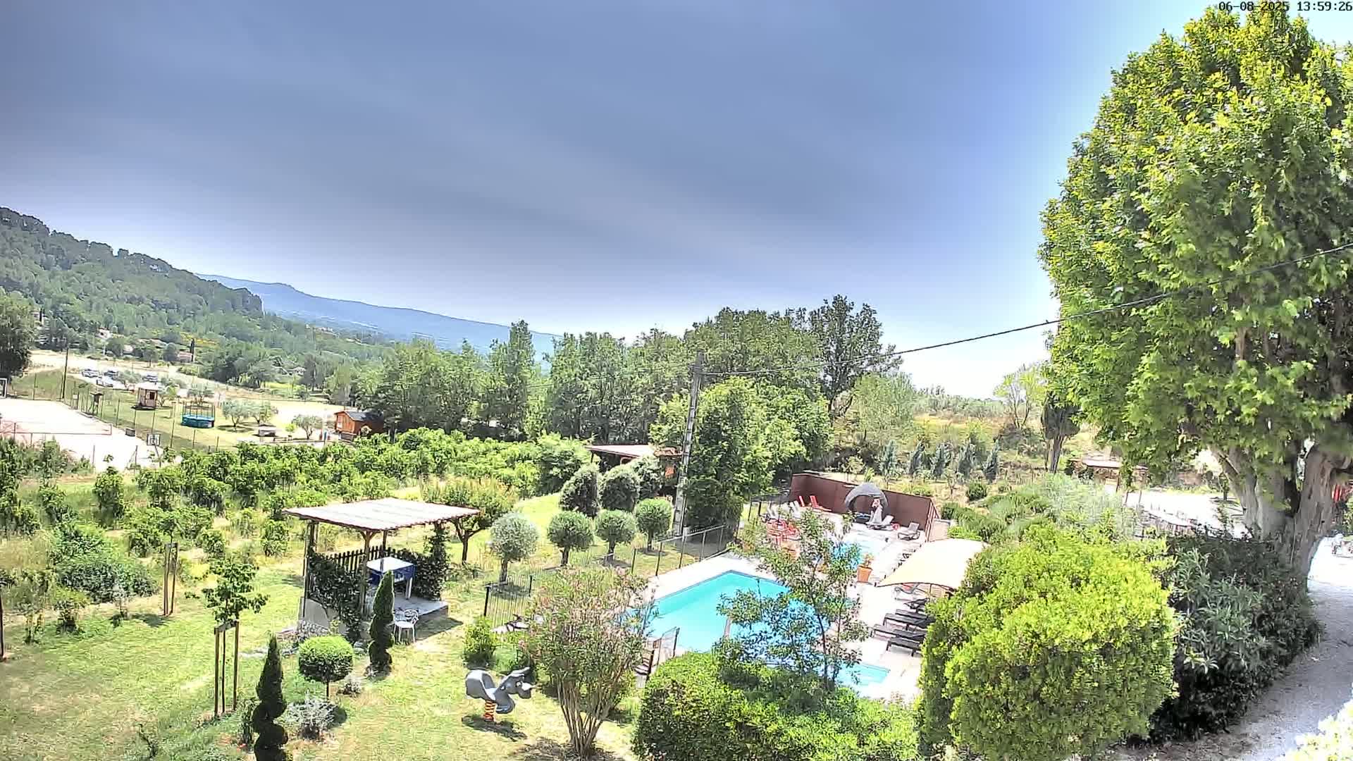 An aerial view of a lush green garden with a swimming pool, surrounded by trees and a distant mountain range under a clear, bright sky.