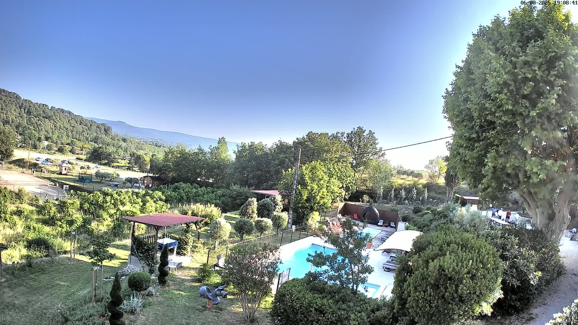 A swimming pool is nestled in a lush green garden with a view of distant mountains under a clear blue sky.