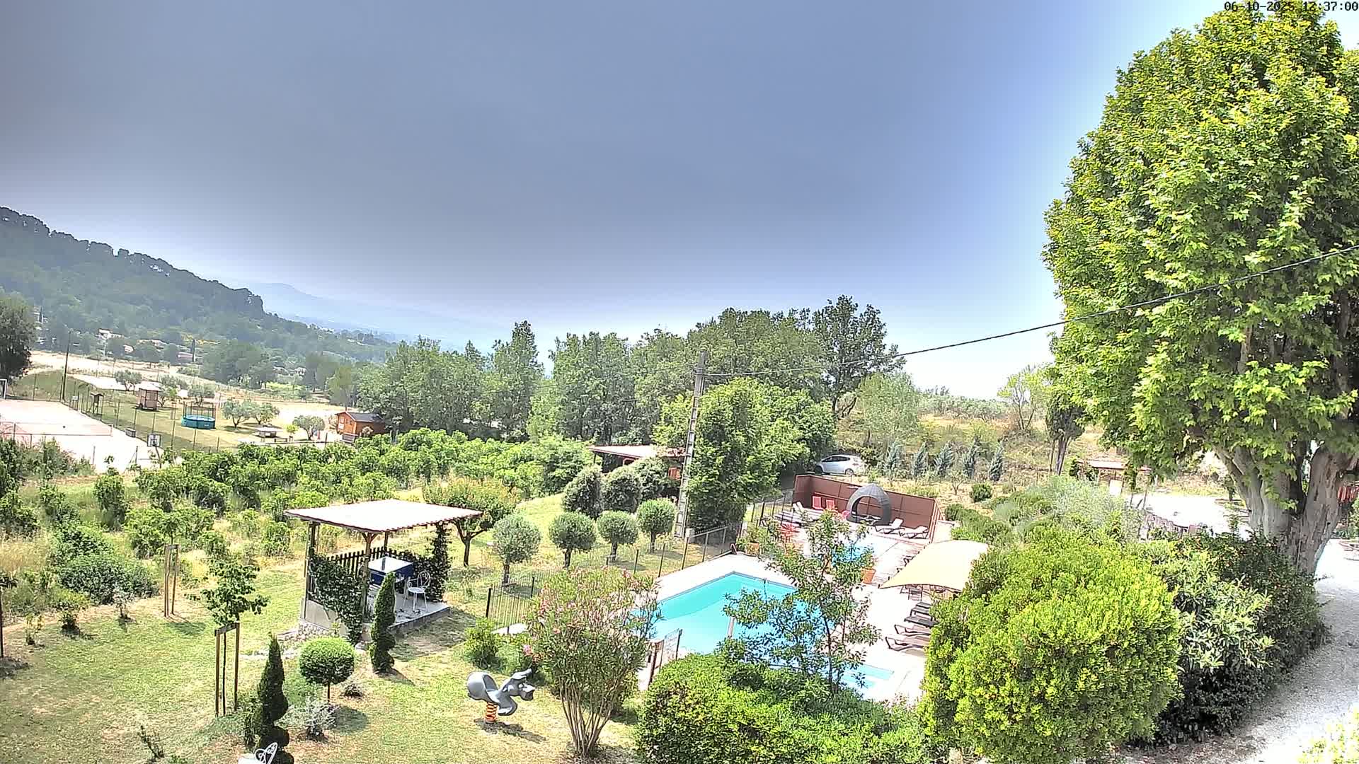 A swimming pool sits in a lush green garden overlooking rolling hills under a clear, mostly sunny sky.