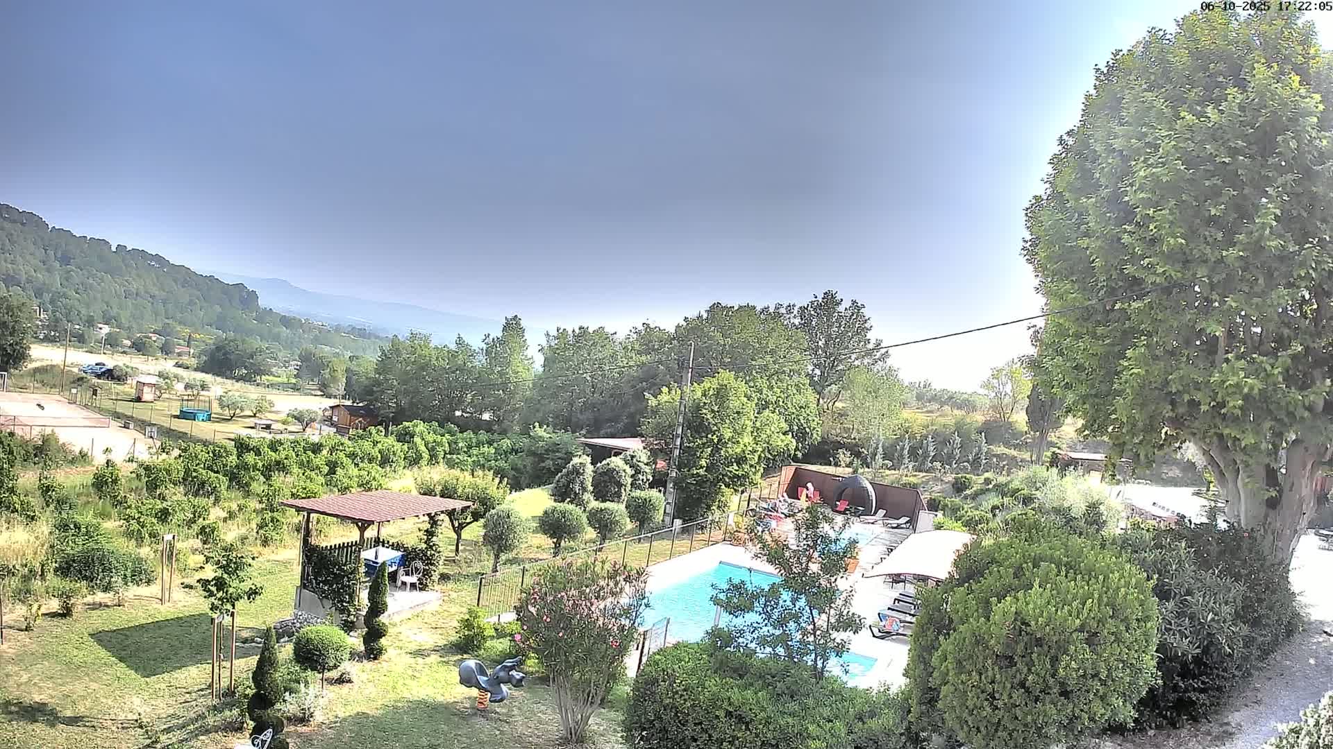 A swimming pool is situated in a lush green garden overlooking a hilly landscape under a clear, bright sky.