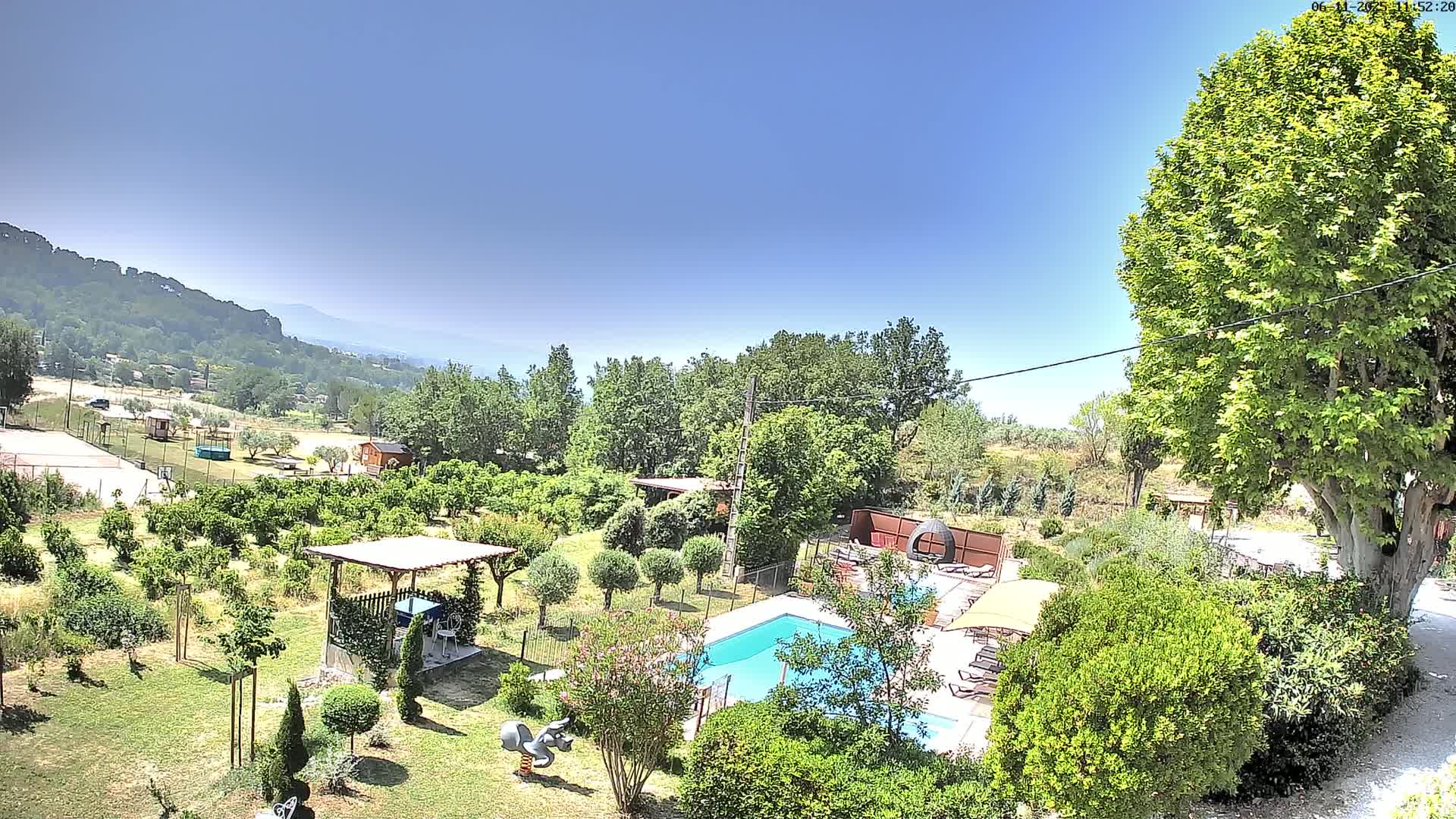 An aerial view shows a swimming pool surrounded by lush green landscaping, a pergola, and several trees under a clear blue sky.