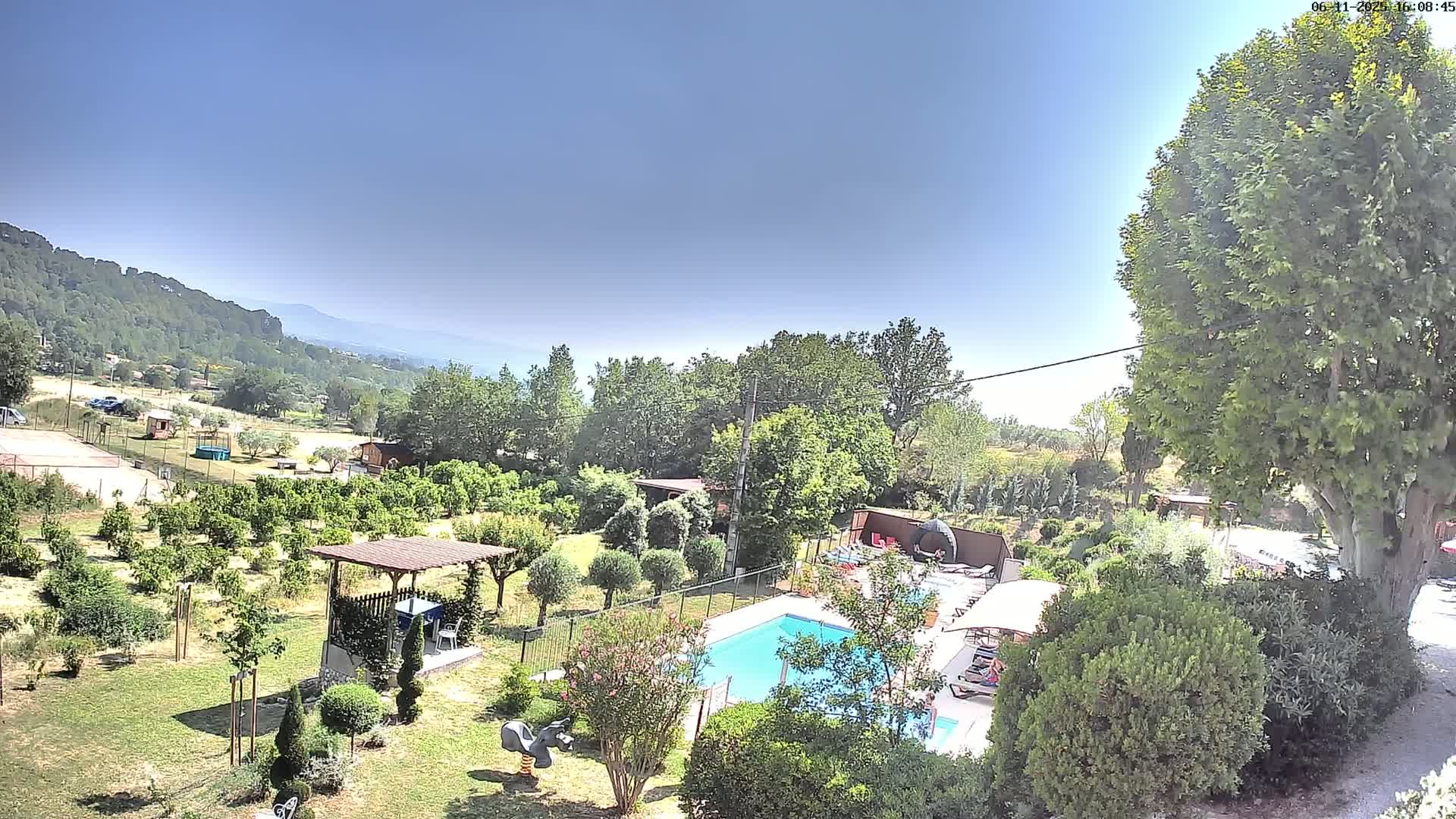 An aerial view shows a swimming pool surrounded by lush greenery, a pergola, and other outdoor features, all under a clear, sunny sky with distant mountains visible.