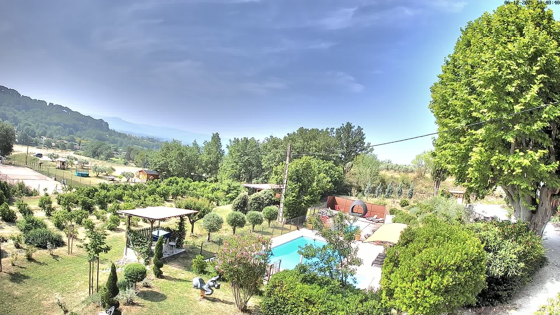 An aerial view shows a swimming pool nestled in a lush green garden with a pergola, fruit trees, and a backdrop of hills under a mostly clear, sunny sky.