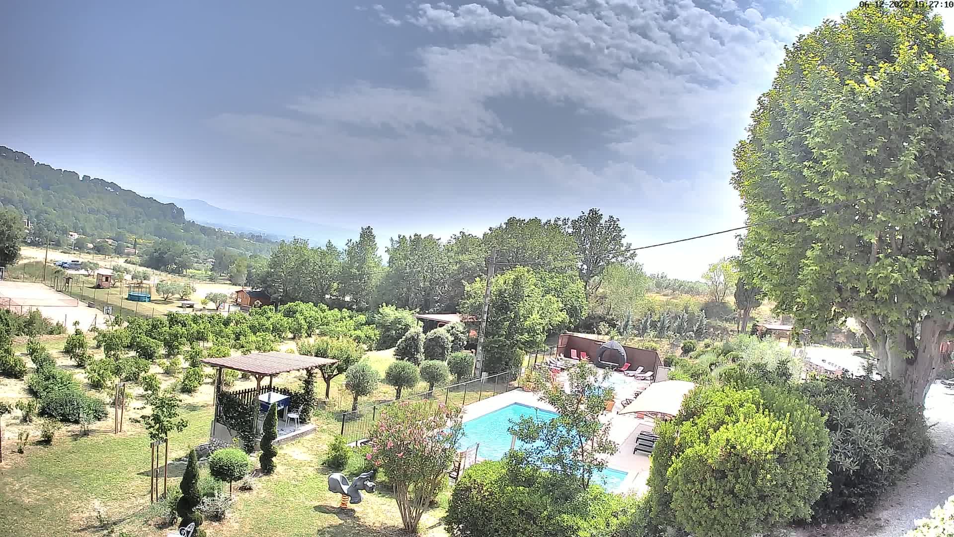 A swimming pool is situated in a lush green garden with a variety of trees and shrubs, overlooking a hilly landscape under a partly cloudy sky.