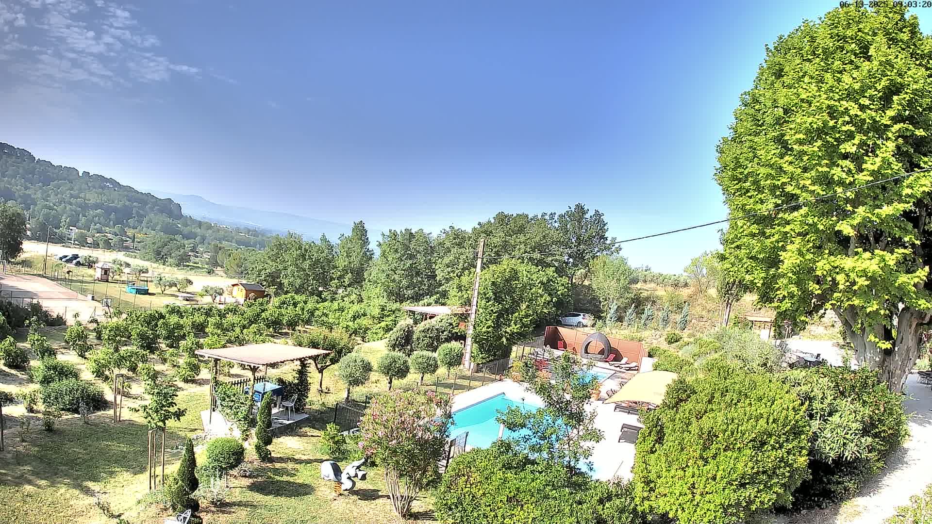 A swimming pool and lush garden are nestled in a rural setting under a clear blue sky, with distant mountains visible in the background.