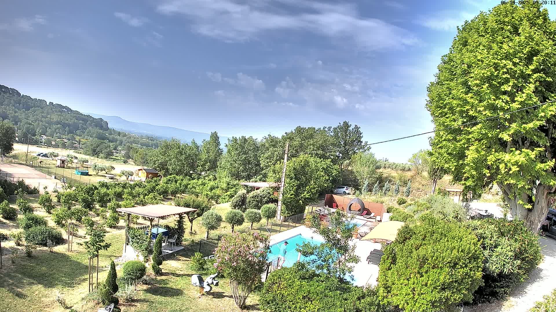 A partly cloudy day reveals a lush, green backyard with a swimming pool, a person standing near it, and a view of distant hills and mountains.
