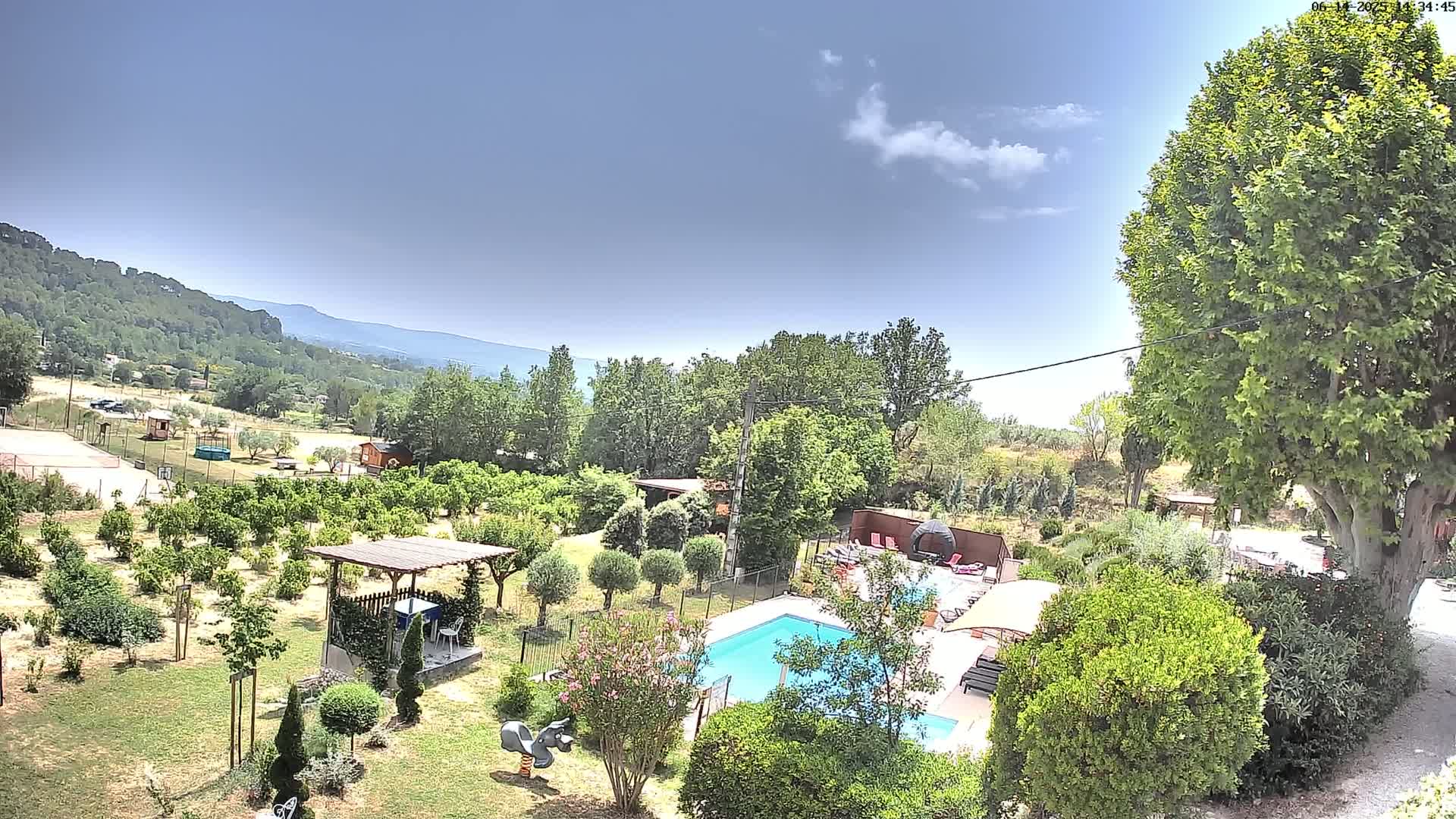 A swimming pool is nestled in a lush green garden, overlooking a hilly landscape under a mostly sunny sky.