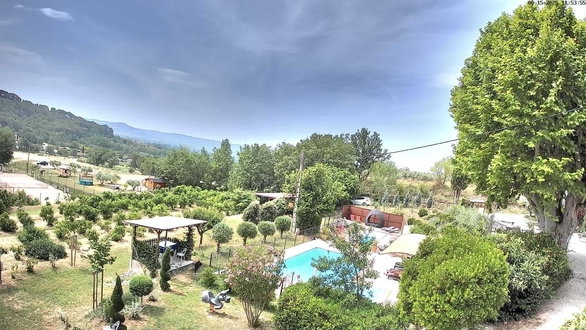 A swimming pool and surrounding garden sit in a rural setting under a partly cloudy sky, with a mountain range visible in the distance.