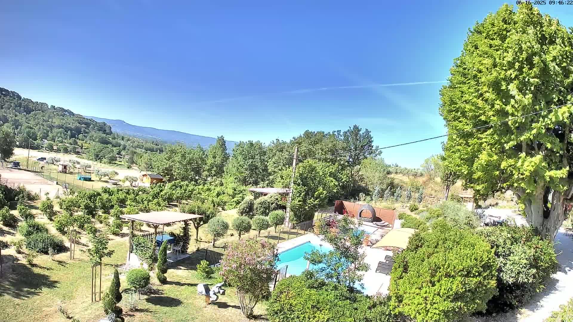 An aerial view shows a sunny day overlooking a backyard with a swimming pool, a pergola, lush greenery, and trees, all set against a backdrop of distant mountains.