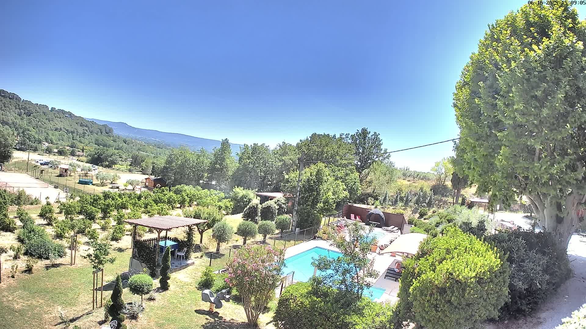 A swimming pool sits in a lush, green landscape with a clear blue sky, overlooking a mountainous region.