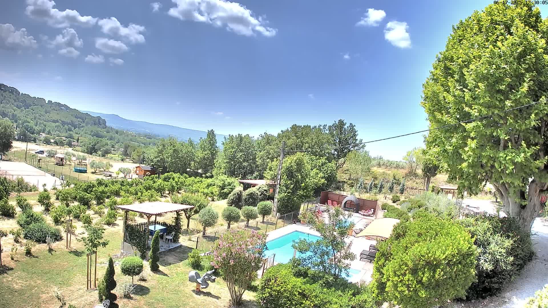 An aerial view of a swimming pool and surrounding garden in a sunny, partly cloudy day, with mountains visible in the background.
