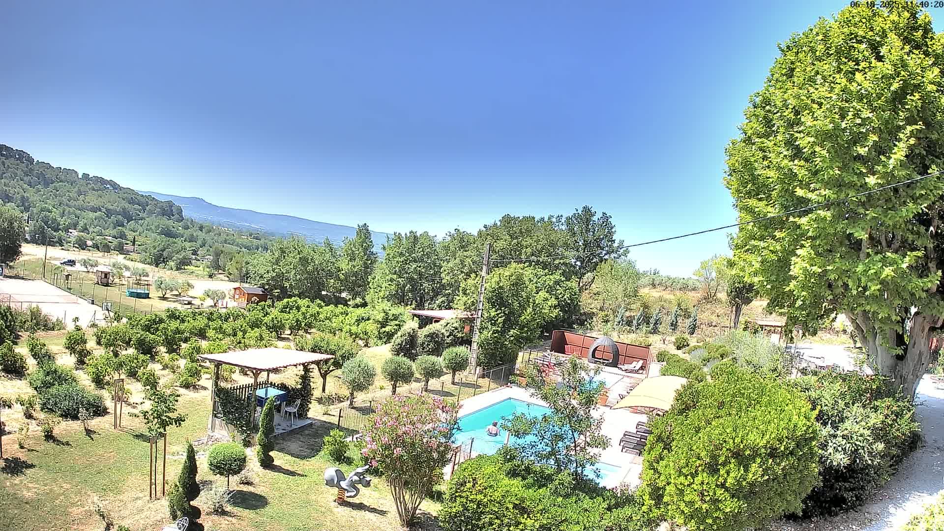 A swimming pool and a lush garden, surrounded by trees and hills under a clear blue sky.