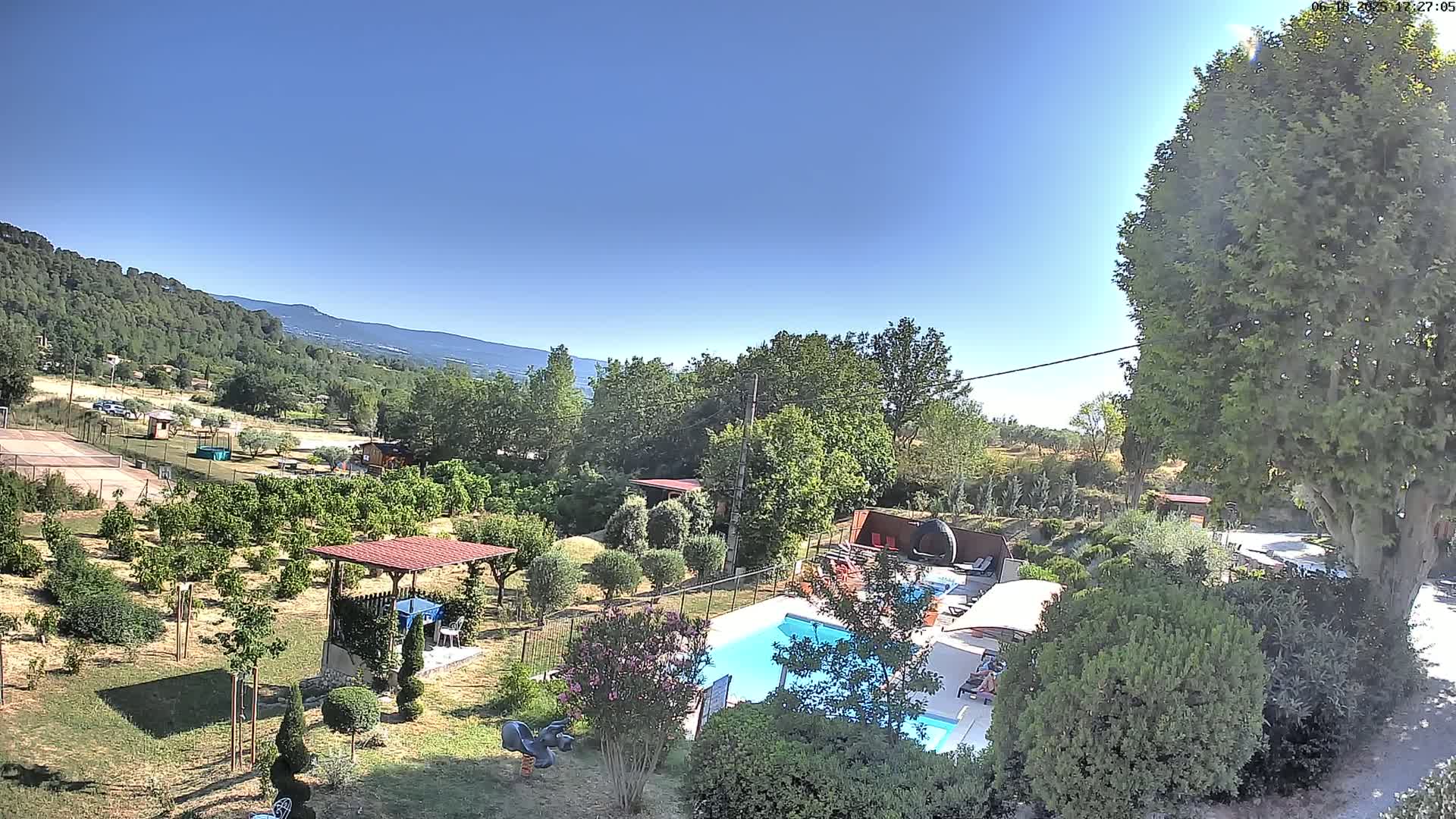 An aerial view shows a swimming pool surrounded by a lush, green garden with various trees and shrubs, a pavilion, and playground equipment, all under a clear, sunny sky and backed by distant hills.