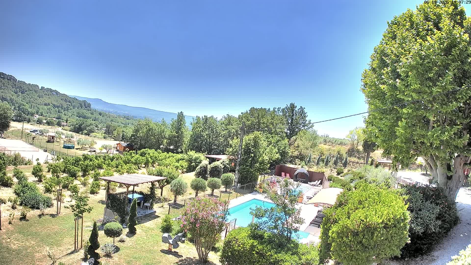 A swimming pool surrounded by lush greenery and trees sits beneath a clear blue sky, overlooking a hilly landscape in the distance.