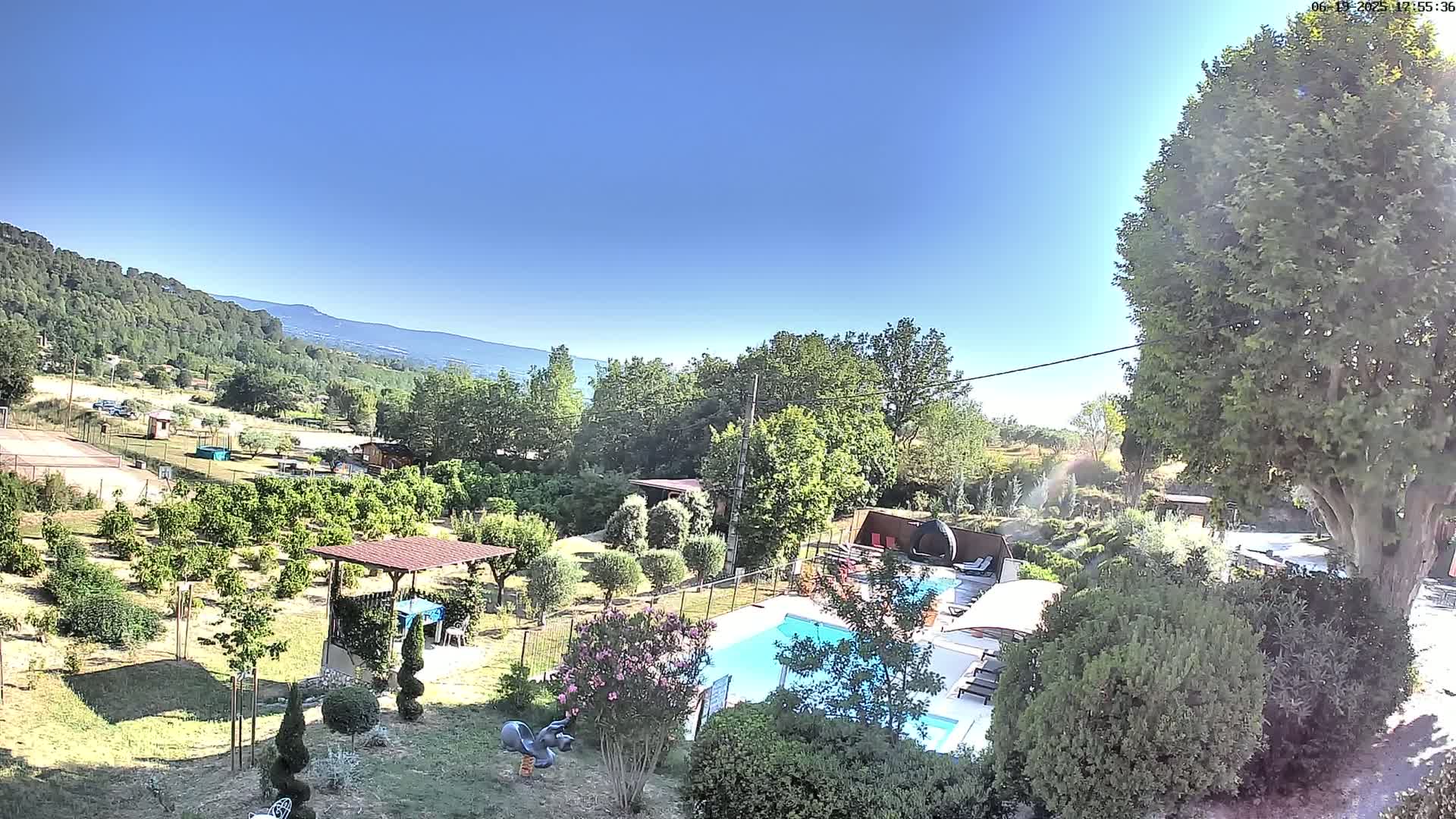 A swimming pool sits in a lush, green garden with a view of distant mountains under a clear blue sky.