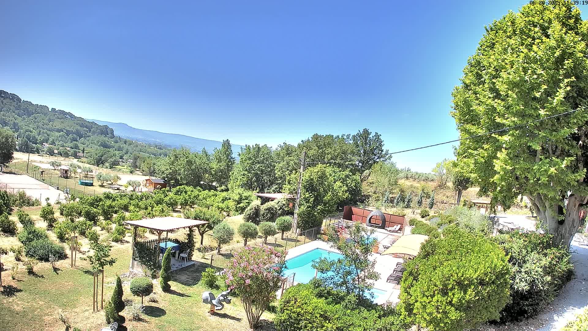 A swimming pool sits in a lush green garden with fruit trees, a pergola, and a view of distant mountains under a clear blue sky.