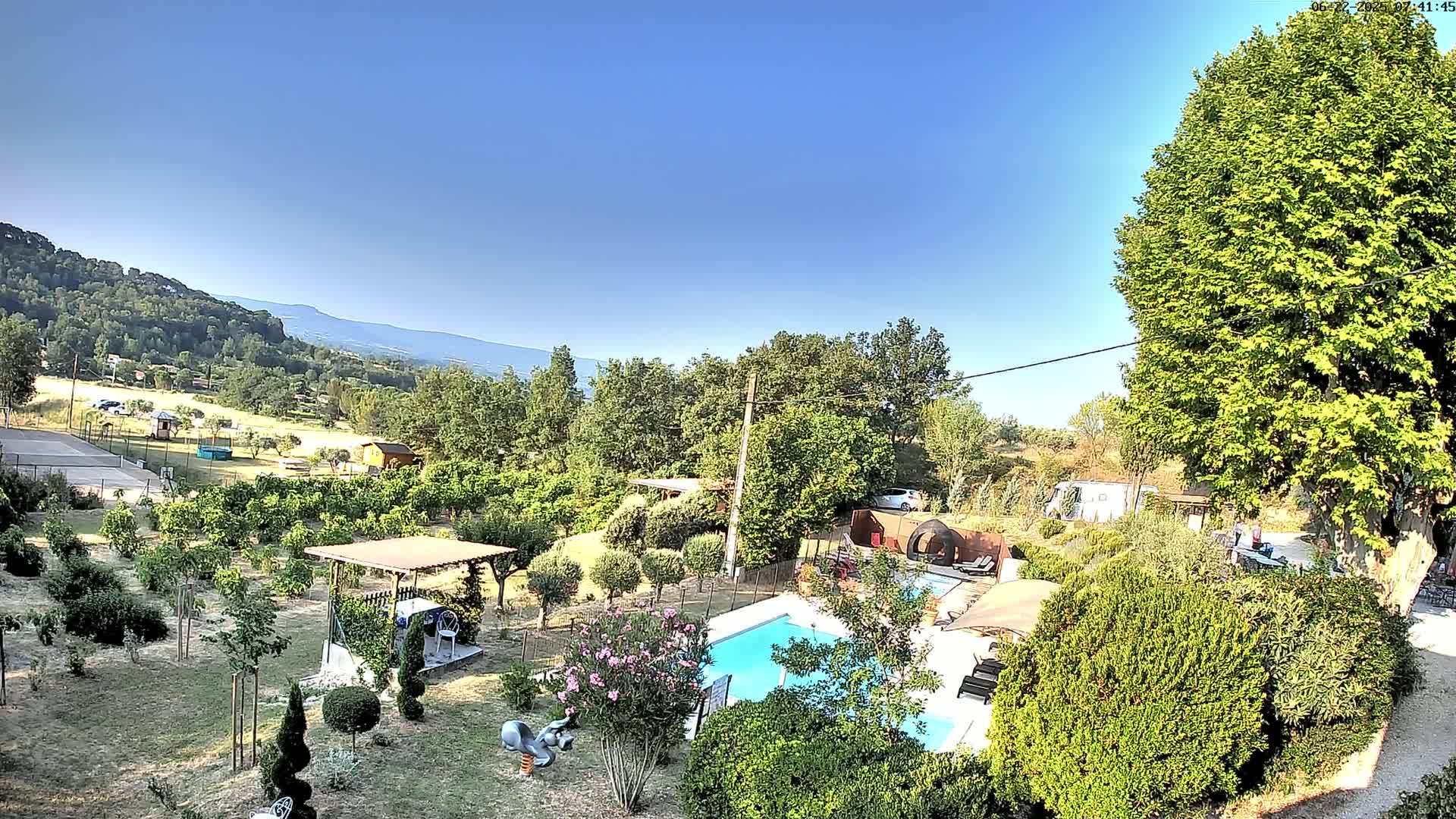 An aerial view of a lush garden with a swimming pool, surrounded by trees and shrubs, on a clear, sunny day with a mountain range in the distance.