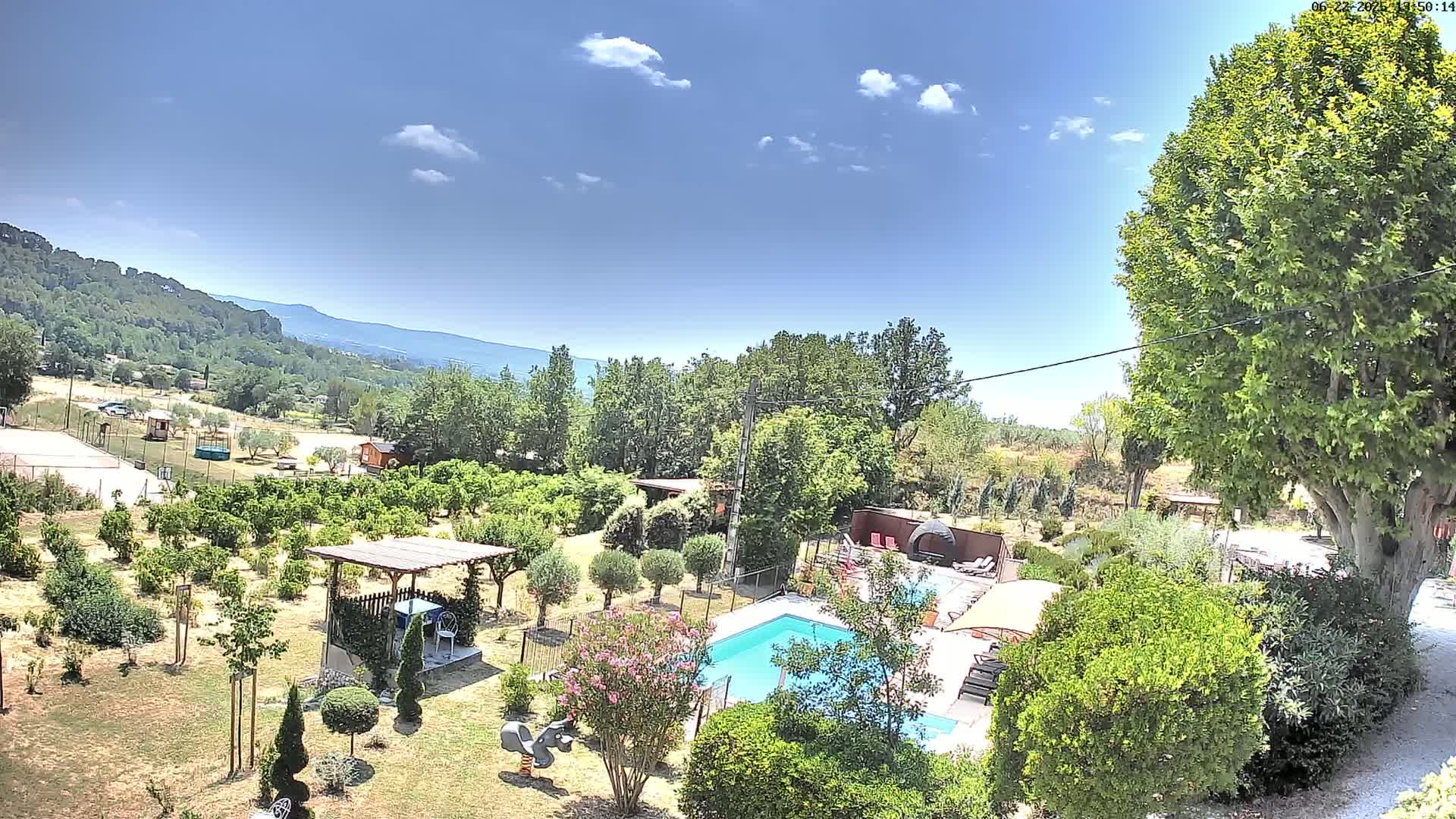 An aerial view of a sunny, landscaped property with a swimming pool, numerous trees and shrubs, and a pergola, overlooking a distant mountain range.