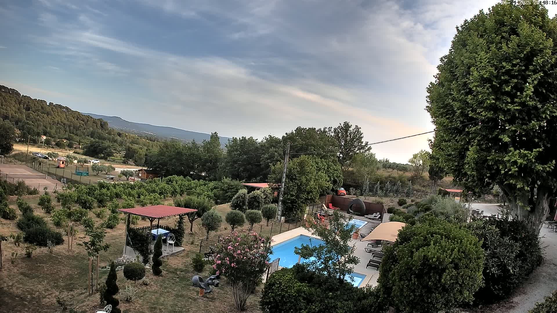 An elevated view shows a swimming pool and surrounding garden with a pergola and various shrubs and trees, under a partly cloudy sky, overlooking a landscape of hills and greenery.