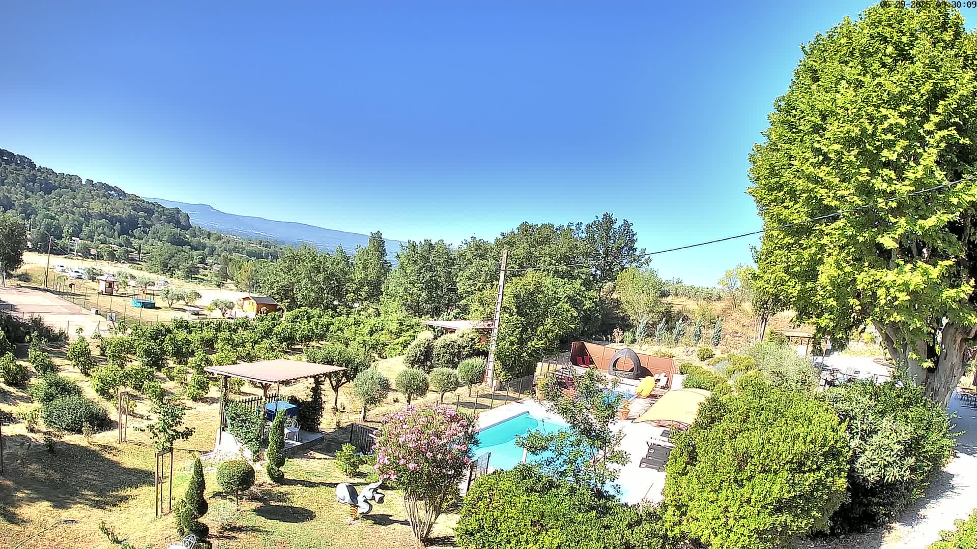 An aerial view shows a swimming pool surrounded by lush greenery and trees, under a clear blue sky, with distant mountains visible in the background.