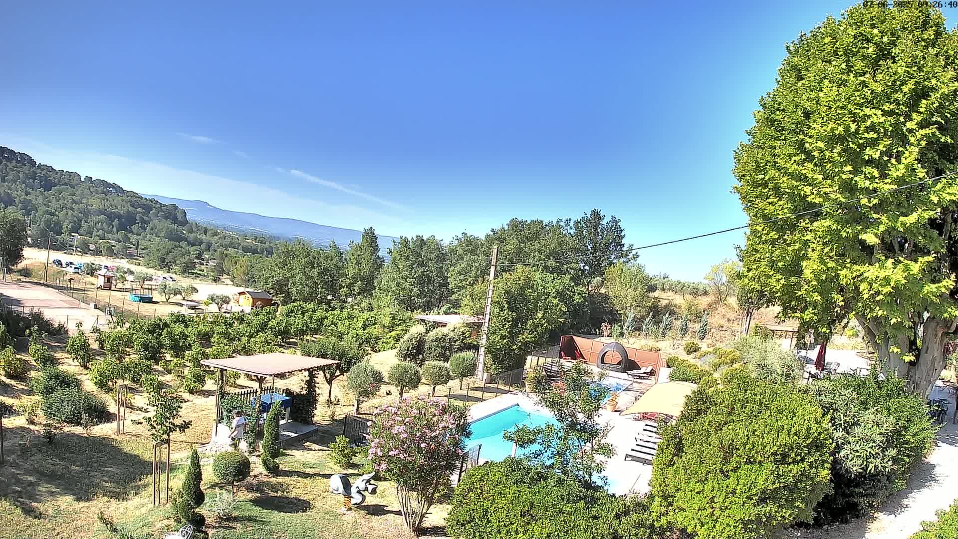 A swimming pool sits in a lush, green garden overlooking a mountain range under a clear, sunny sky.