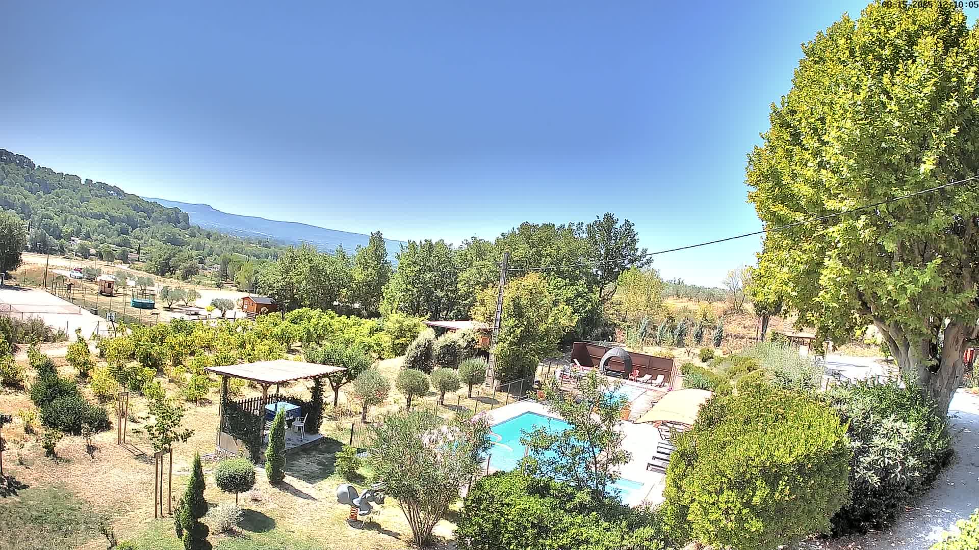 A swimming pool sits in a lush, green garden overlooking a mountain range under a clear, sunny sky.