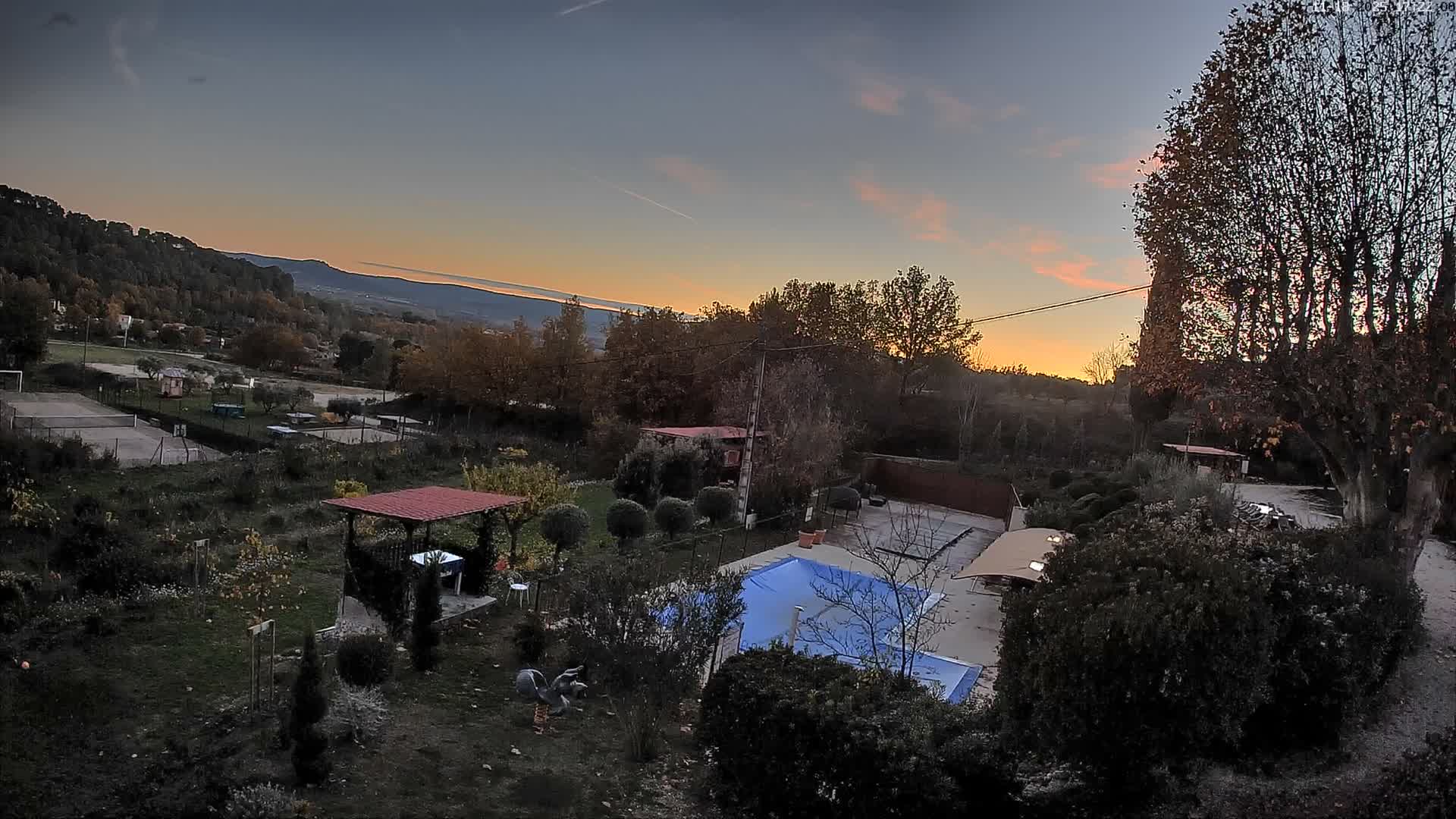 A swimming pool sits in a lush, green garden overlooking a mountain range under a clear, sunny sky.