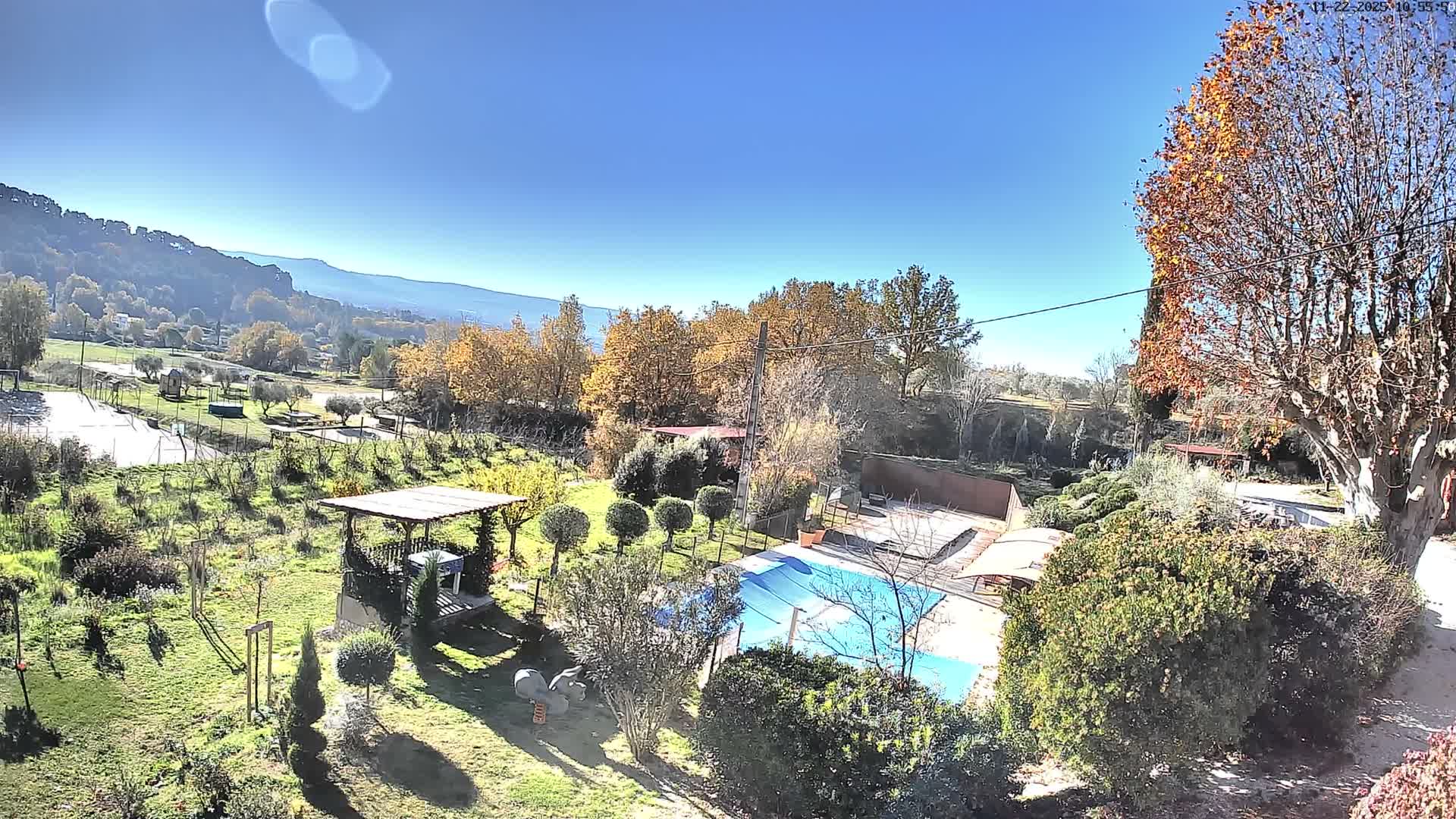 A bright, sunny autumn day illuminates a diverse outdoor landscape featuring a blue swimming pool with a surrounding deck, a grassy garden area with a wooden gazebo and play equipment, and distant mountains under a clear blue sky, all dotted with trees displaying fall foliage.