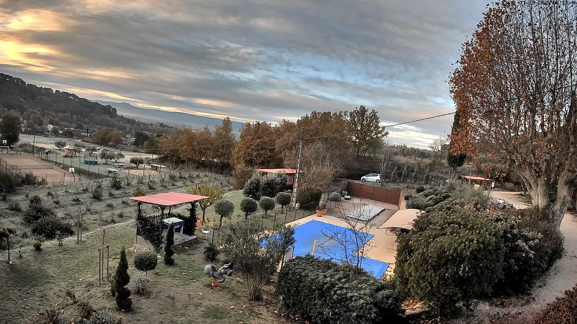 A sprawling, frost-covered rural landscape unfolds under a dramatically cloudy sky with hints of golden light, featuring a blue-covered swimming pool, a gazebo, deciduous trees, a tennis court, and distant hills, all suggesting a cold morning.