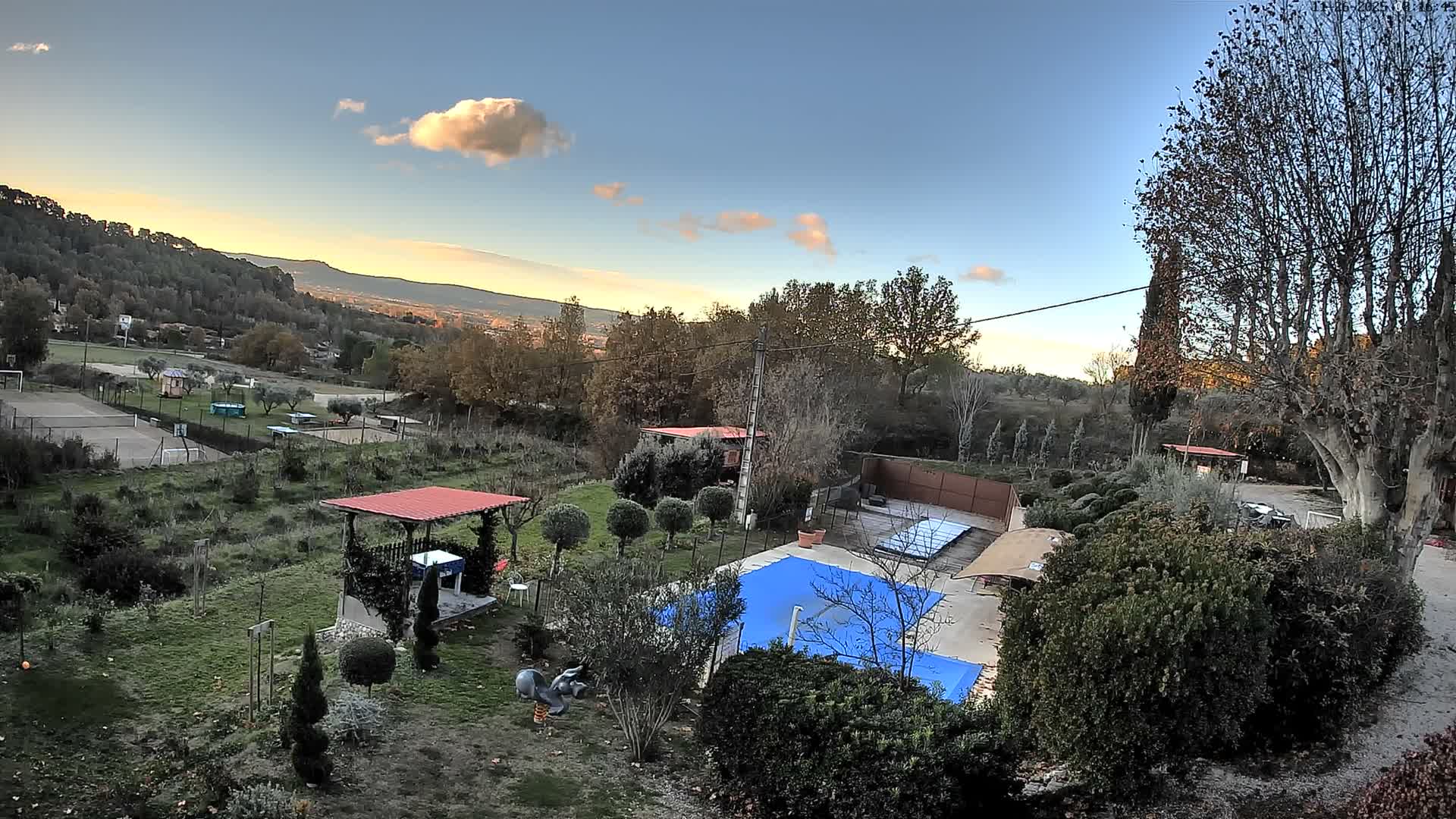 A panoramic outdoor scene reveals a landscaped garden with a blue-covered swimming pool and a red-roofed gazebo in the foreground, stretching towards distant sports courts, buildings, and tree-covered hills, all under a clear sky with scattered clouds and a warm golden glow near the horizon, indicating late afternoon.