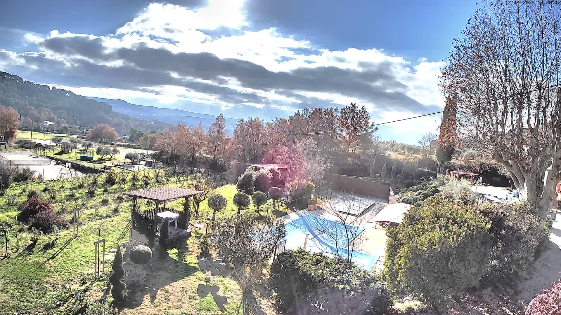 The image captures a broad outdoor scene of a rural property with a blue-covered swimming pool, a red-roofed gazebo, and sports facilities, all set against a backdrop of autumnal hills under a heavy, dark overcast sky.