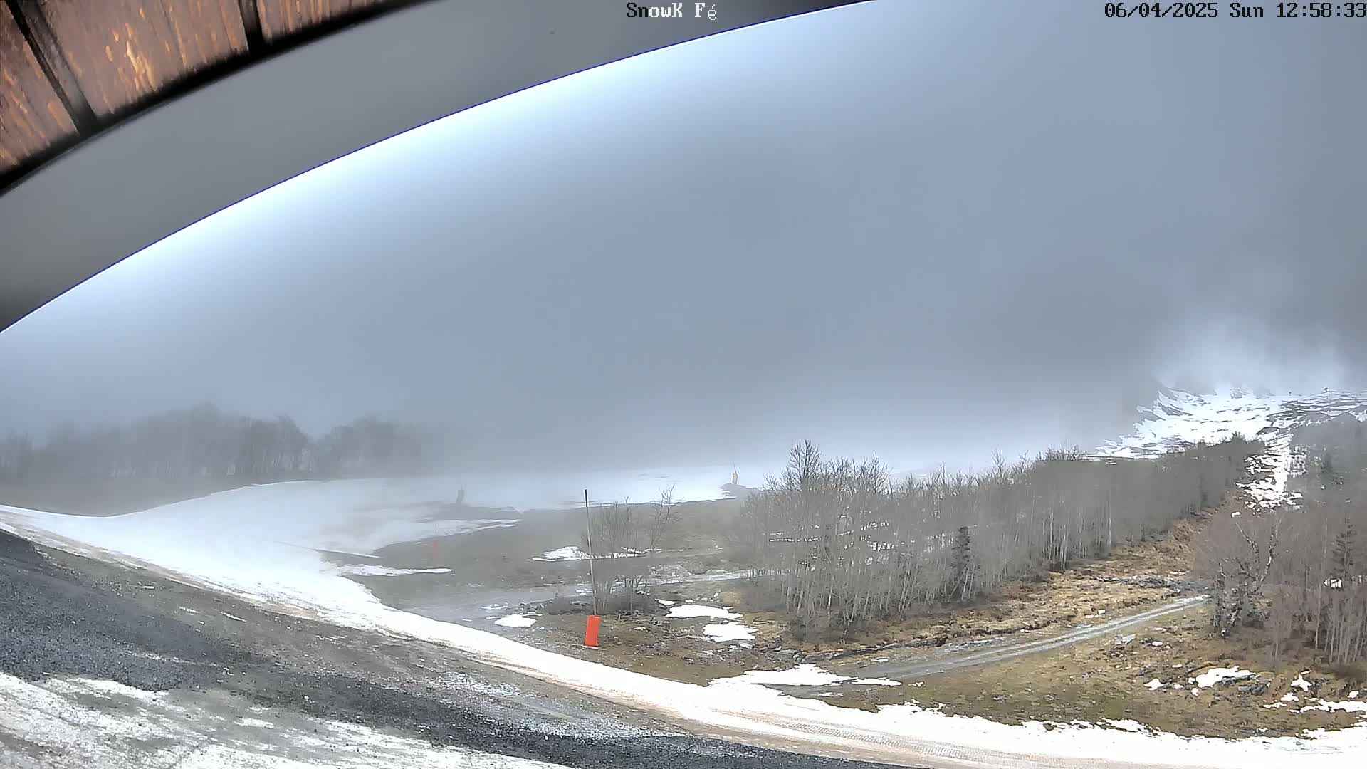 A foggy, snowy landscape is seen from under an overhanging structure, showing a mostly snow-covered area with bare trees and a dirt road.