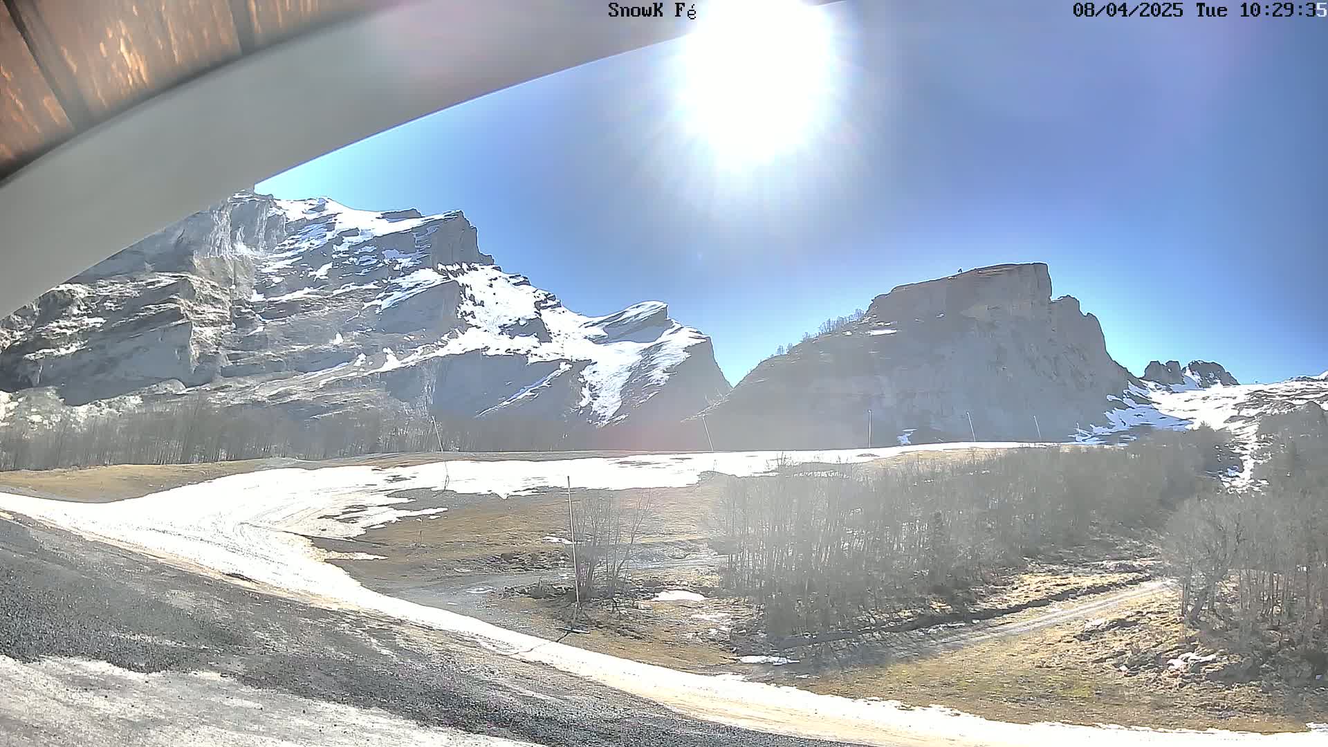 A sunny day reveals snow-capped mountains and a valley with patches of snow and bare trees.