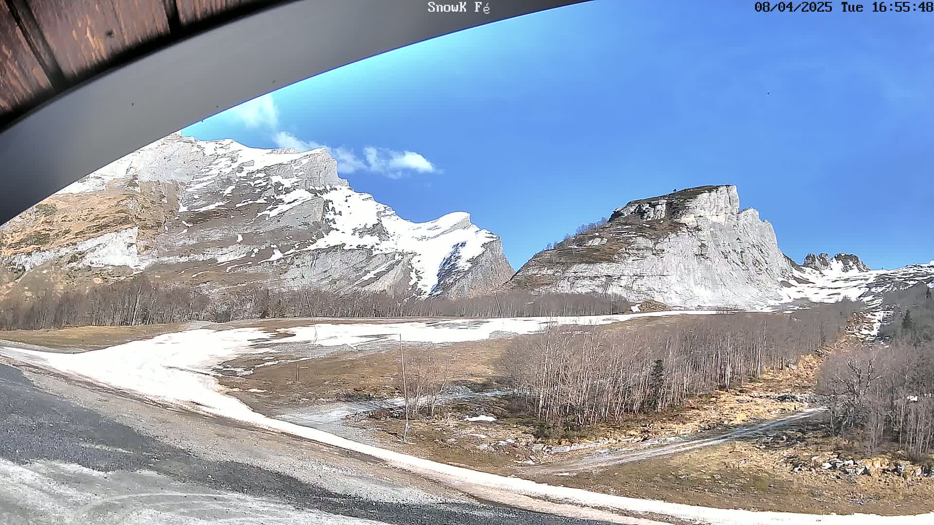 A sunny day reveals snow-capped mountains and a valley with patches of snow and bare trees.