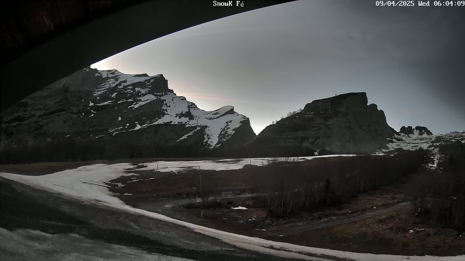 A view from under a structure shows snow-capped mountains under a dusky sky, with patches of snow on the ground in the foreground.