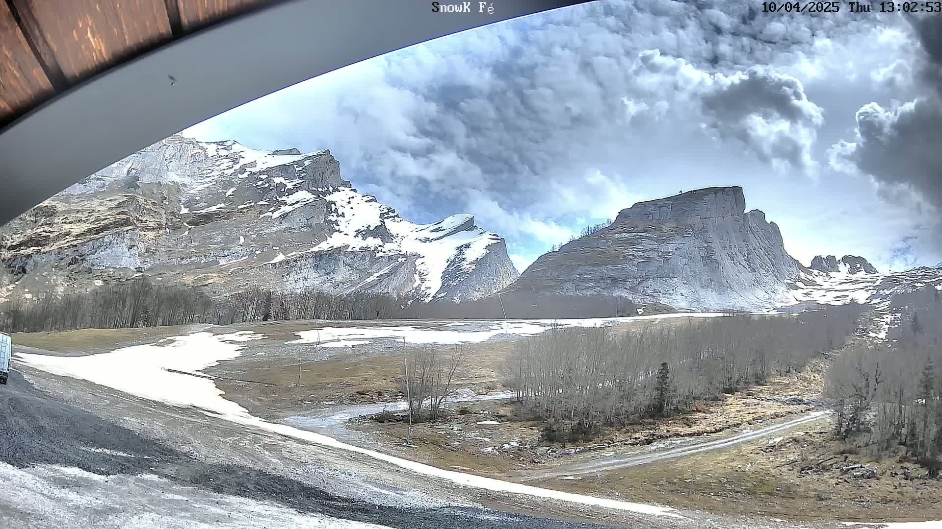 A view from under a wooden overhang shows snow-capped mountains under a partly cloudy sky, with patches of snow and bare trees in the valley below.
